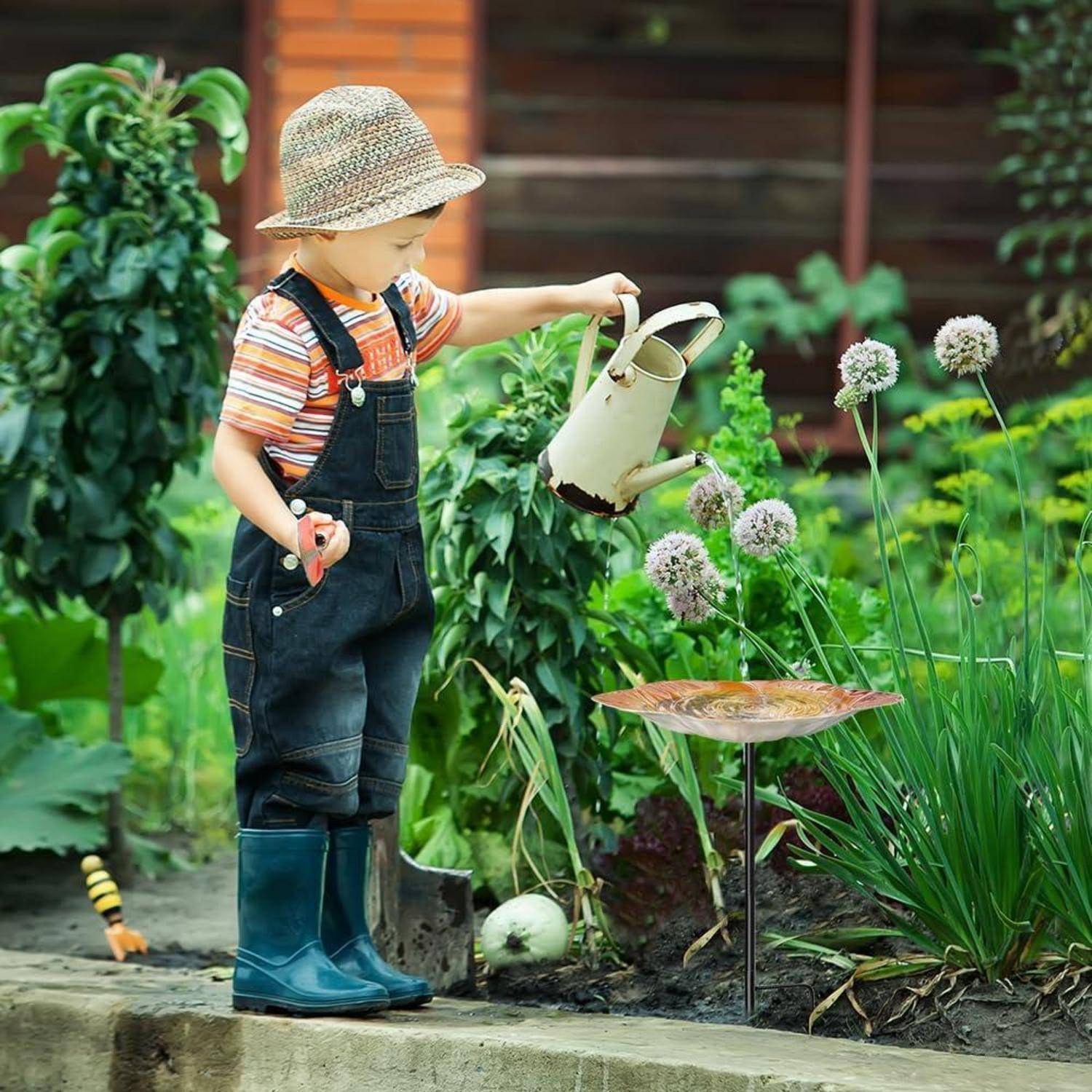 Junges Kind in Latzhose und Stiefeln gießt Gartenpflanzen mit einer Gießkanne neben blühenden Alliumblüten.
