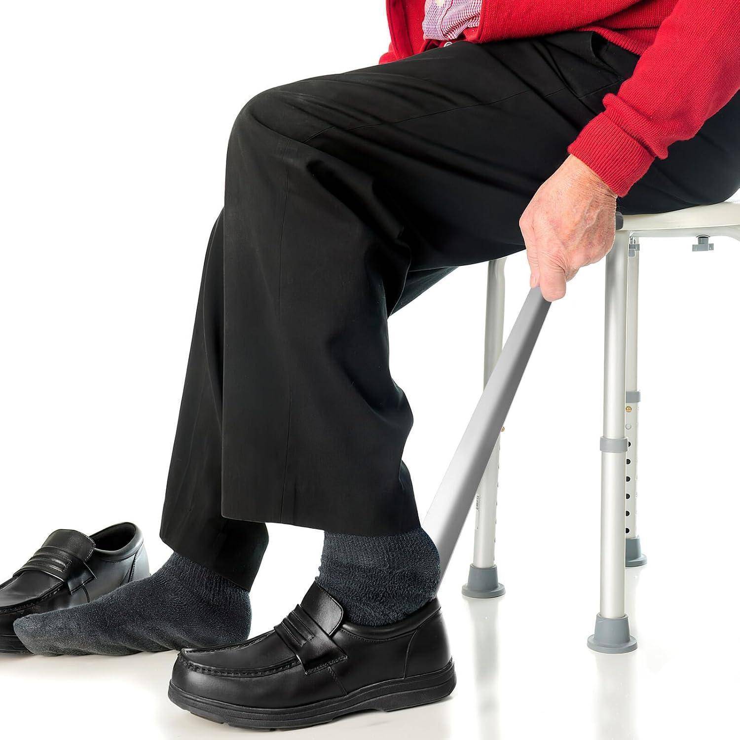 Ältere Person, die auf einem Duschstuhl sitzt und sich mit einer Haltestange beim Aufstehen oder Hochkommen hilft, trägt eine schwarze Hose, Socken und Schuhe.