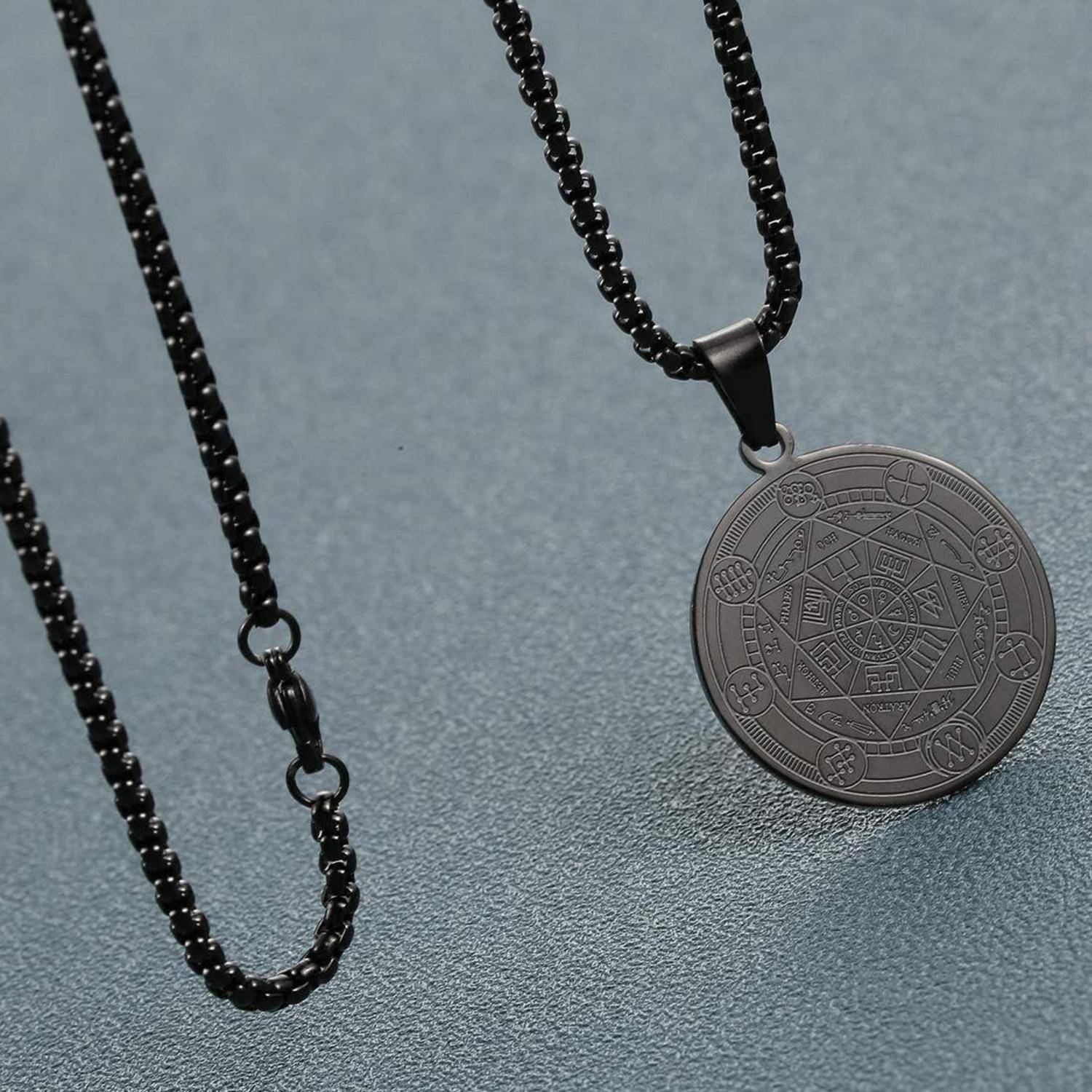 Zwei schwarze Perlenketten auf einer grauen Oberfläche; eine trägt einen runden gravierten Anhänger mit geometrischen und symbolähnlichen Markierungen.