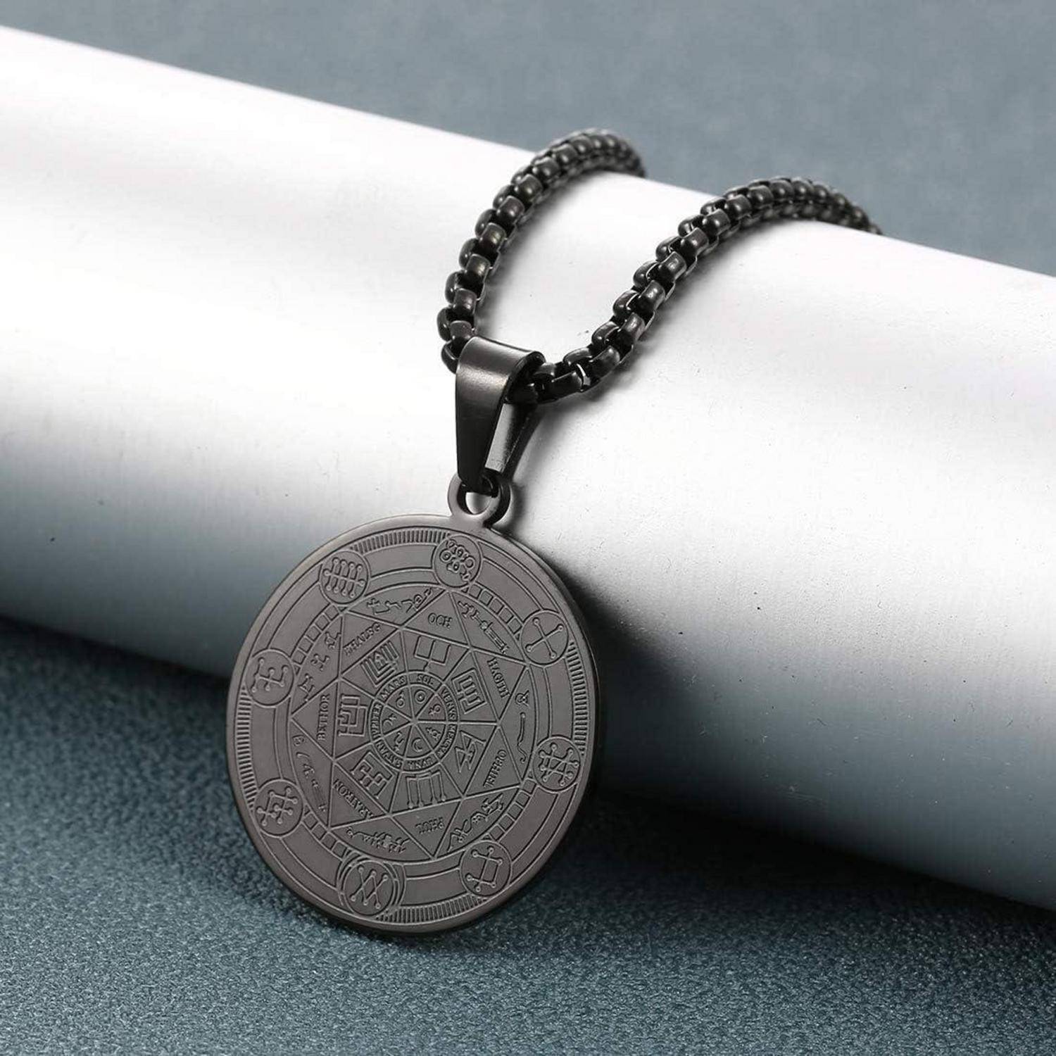 Schwarze Anhängerkette mit einer gravierten kreisförmigen Medaille an einer Kette, die auf einer hellgrauen zylindrischen Oberfläche ruht.