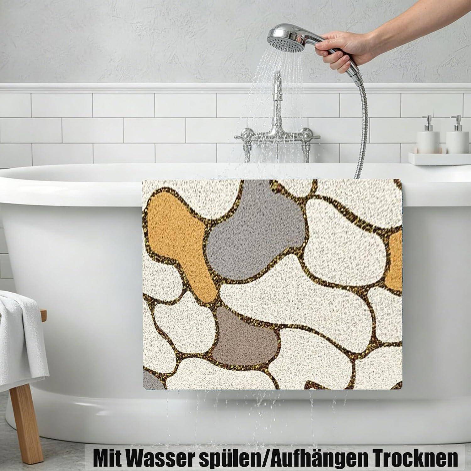 Badematte mit Kieselsteinmuster hängt über einer weißen Badewanne zum Trocknen, während eine Handbrause sie abspült; deutscher Text besagt „Mit Wasser abspülen / zum Trocknen aufhängen