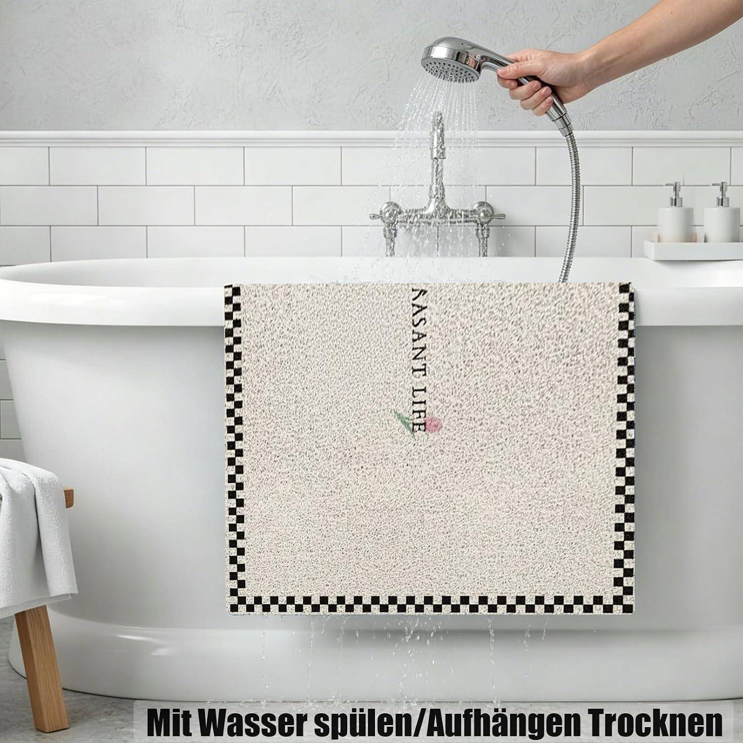 Hand hält einen Duschkopf und spült eine über einer Badewanne drapierte Badmatte aus, mit deutschem Text darunter, der besagt „Mit Wasser spülen/Aufhängen Trocknen