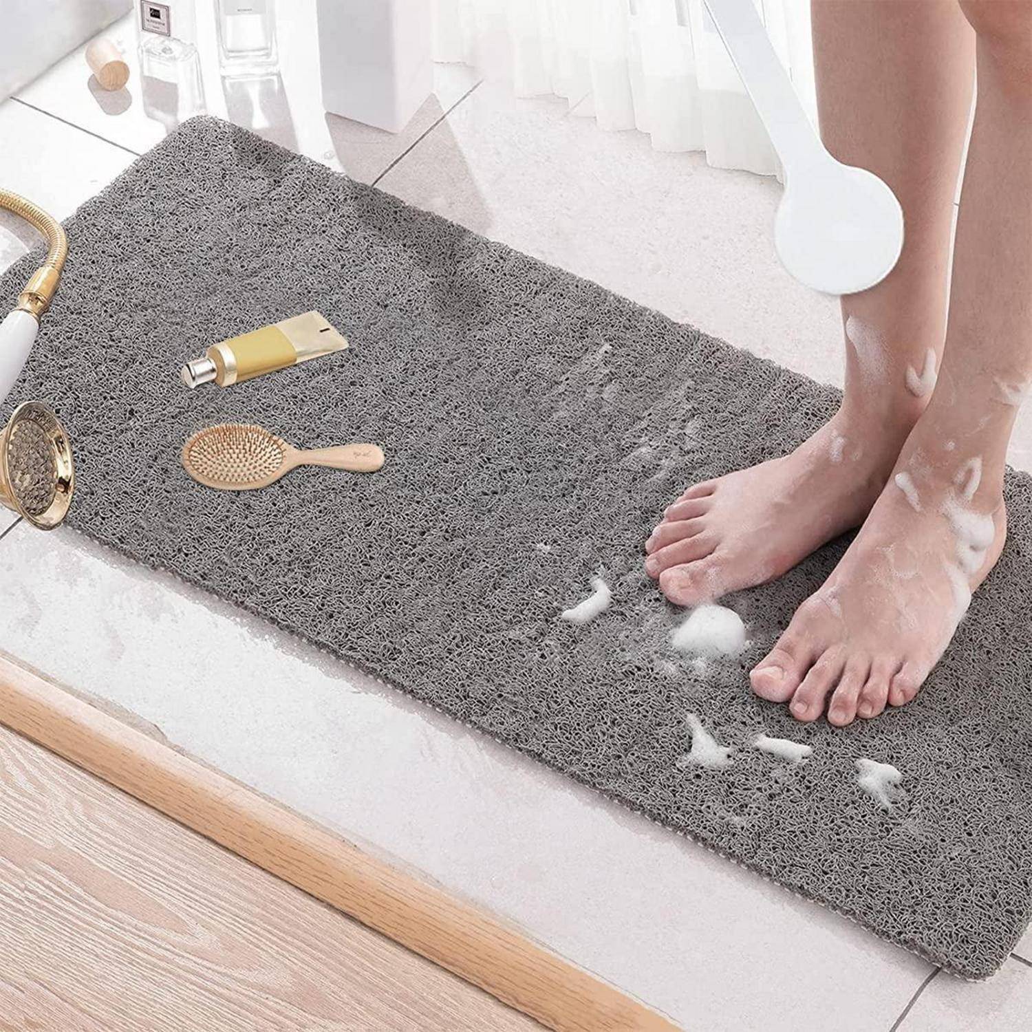 Füße, die auf einer grauen Badematte auf einem Badezimmerboden stehen, mit Badeutensilien in der Nähe, die die Verwendung und Textur der Matte zeigen.