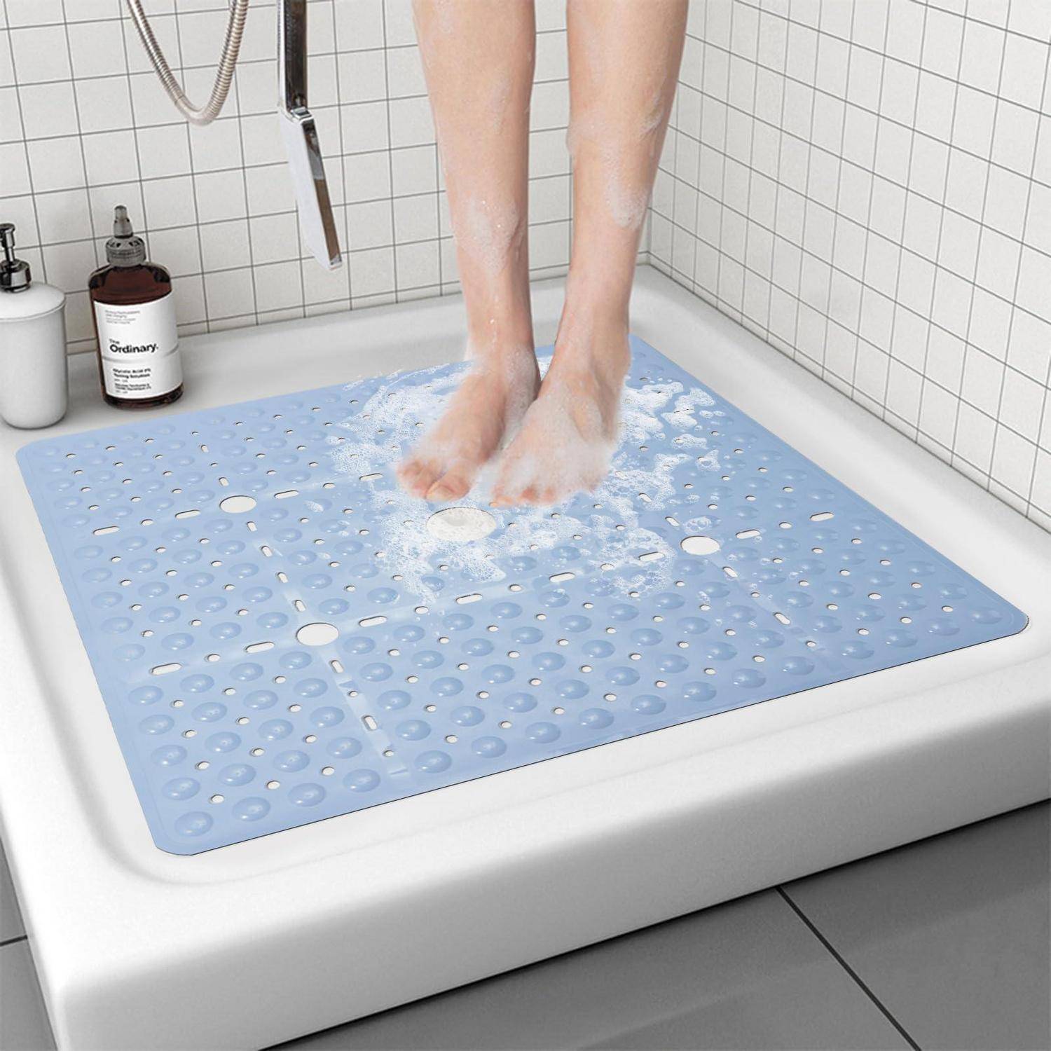 Person steht auf einer blauen strukturierten Badmatte in einer Dusche, mit Seifenschaum um ihre Füße und Badezimmerflaschen in der Nähe.
