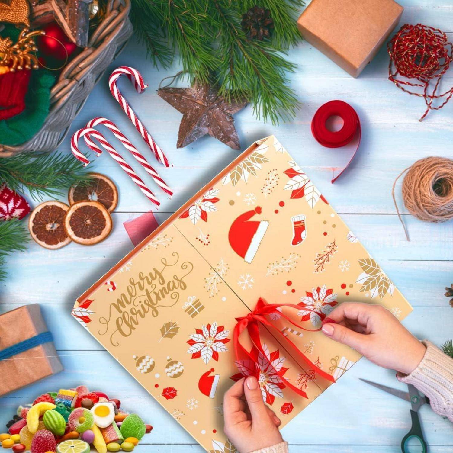 Hände, die ein rotes Band um eine Weihnachtsgeschenkbox binden, umgeben von Zuckerstangen, Christbaumkugeln, getrockneten Zitrusfrüchten und Weihnachtsdeko.