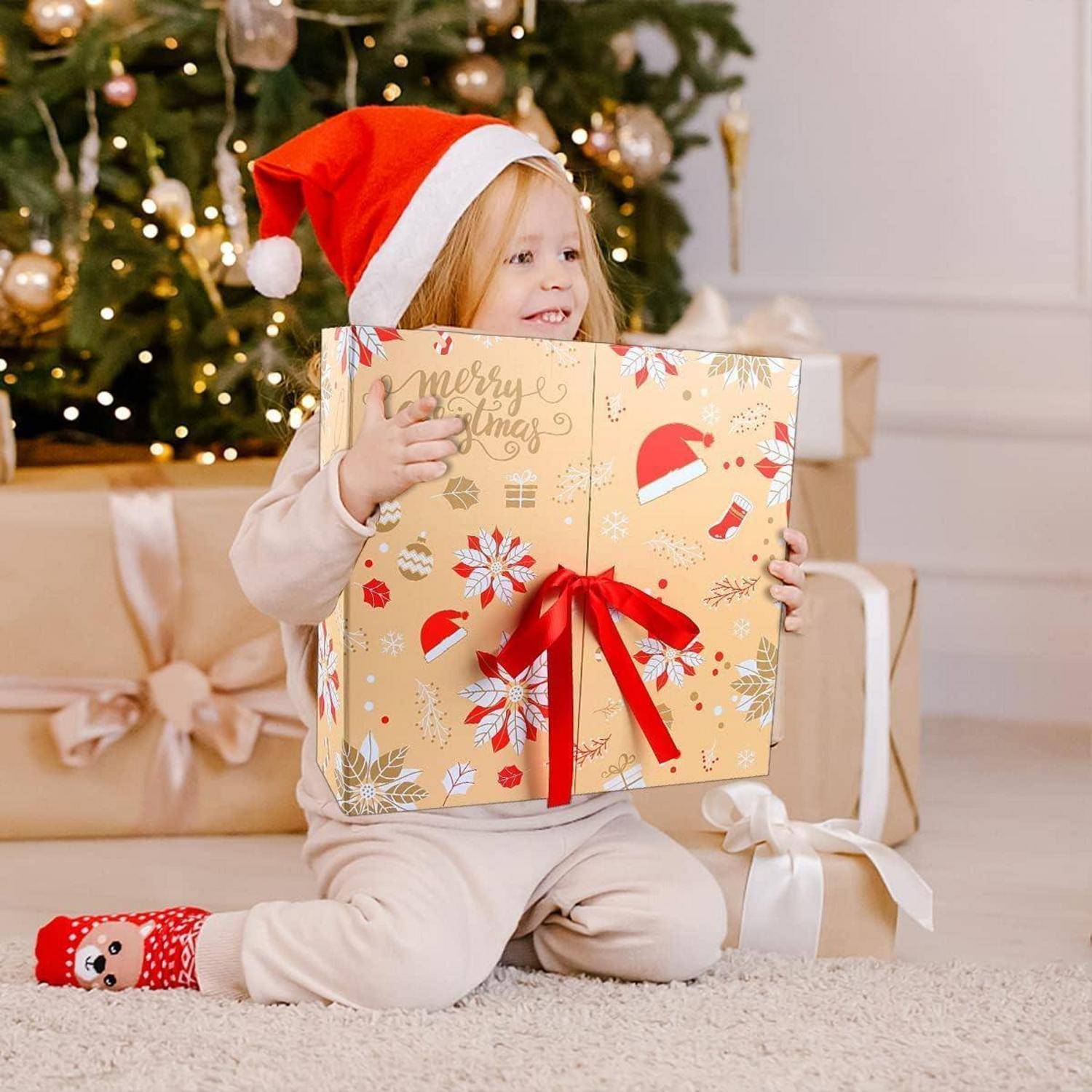Ein junges Kind in einer Weihnachtsmütze sitzt mit einer großen eingepackten Weihnachtsgeschenkbox vor einem geschmückten Baum und Geschenken.