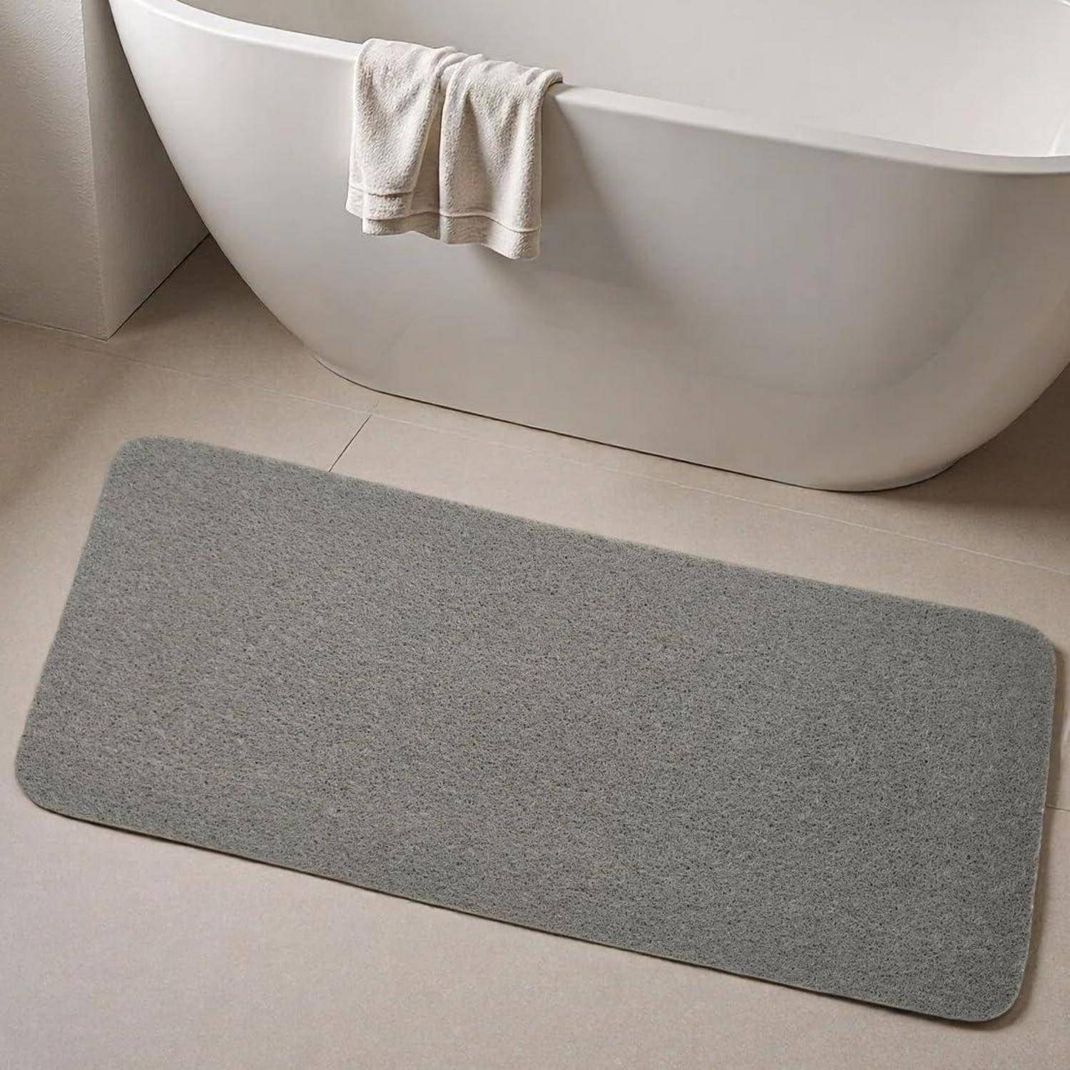 Graue rechteckige Badmatte auf einem beigefarbenem Badezimmerboden neben einer weißen Badewanne mit einem Handtuch, das über den Rand drapiert ist.