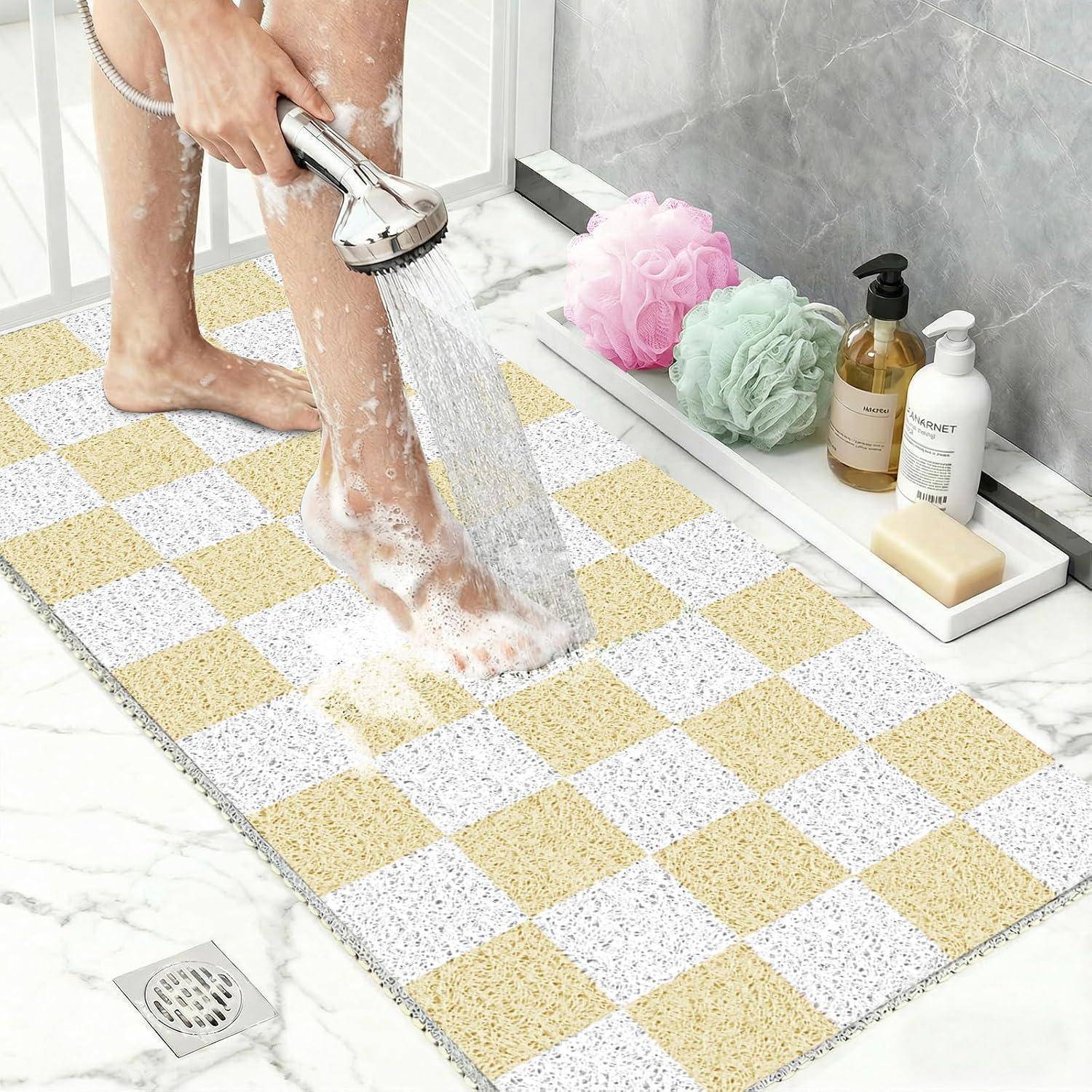 Eine Person spült Seife von einem Bein unter der Dusche ab, mit einer karierten rutschfesten Badmatte und Badeprodukten auf einem nahe gelegenen Regal.