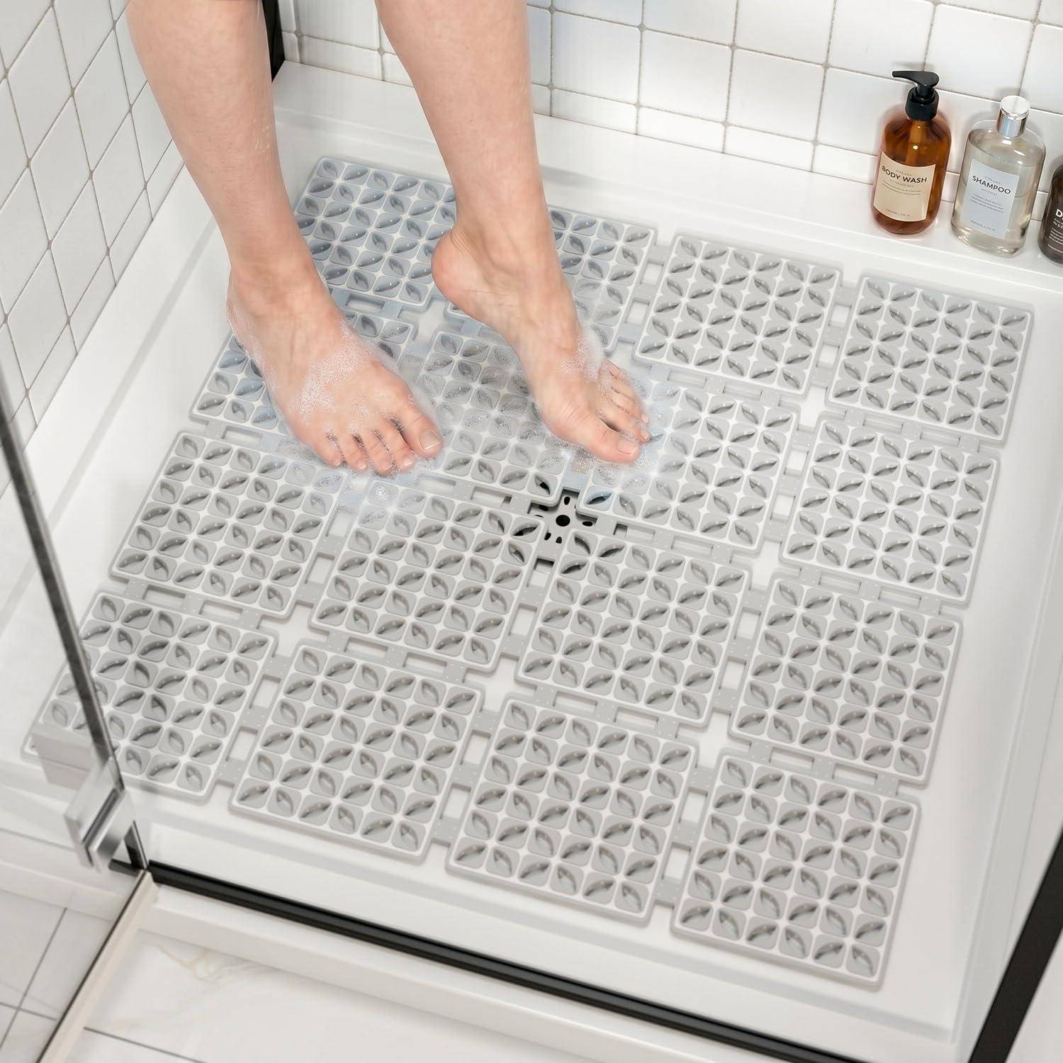 Nackte Füße stehen auf einer grauen rutschfesten Duschenmatte in einer weiß gefliesten Dusche, mit Seifenflaschen auf einem Regal.