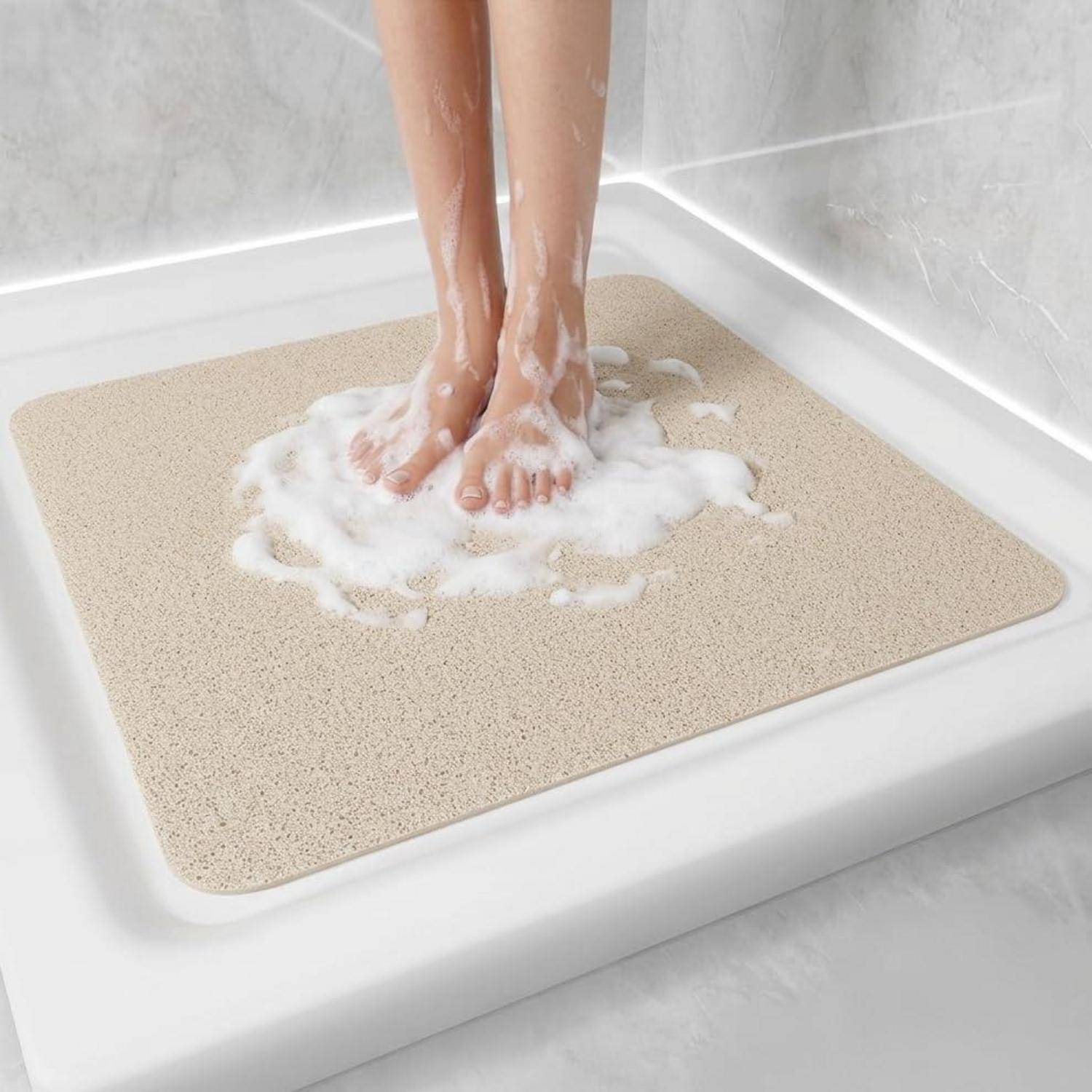 Person, die auf einer beigen rutschfesten Duschmatte in einer weißen Dusche steht, Füße mit Seifenschaum bedeckt.