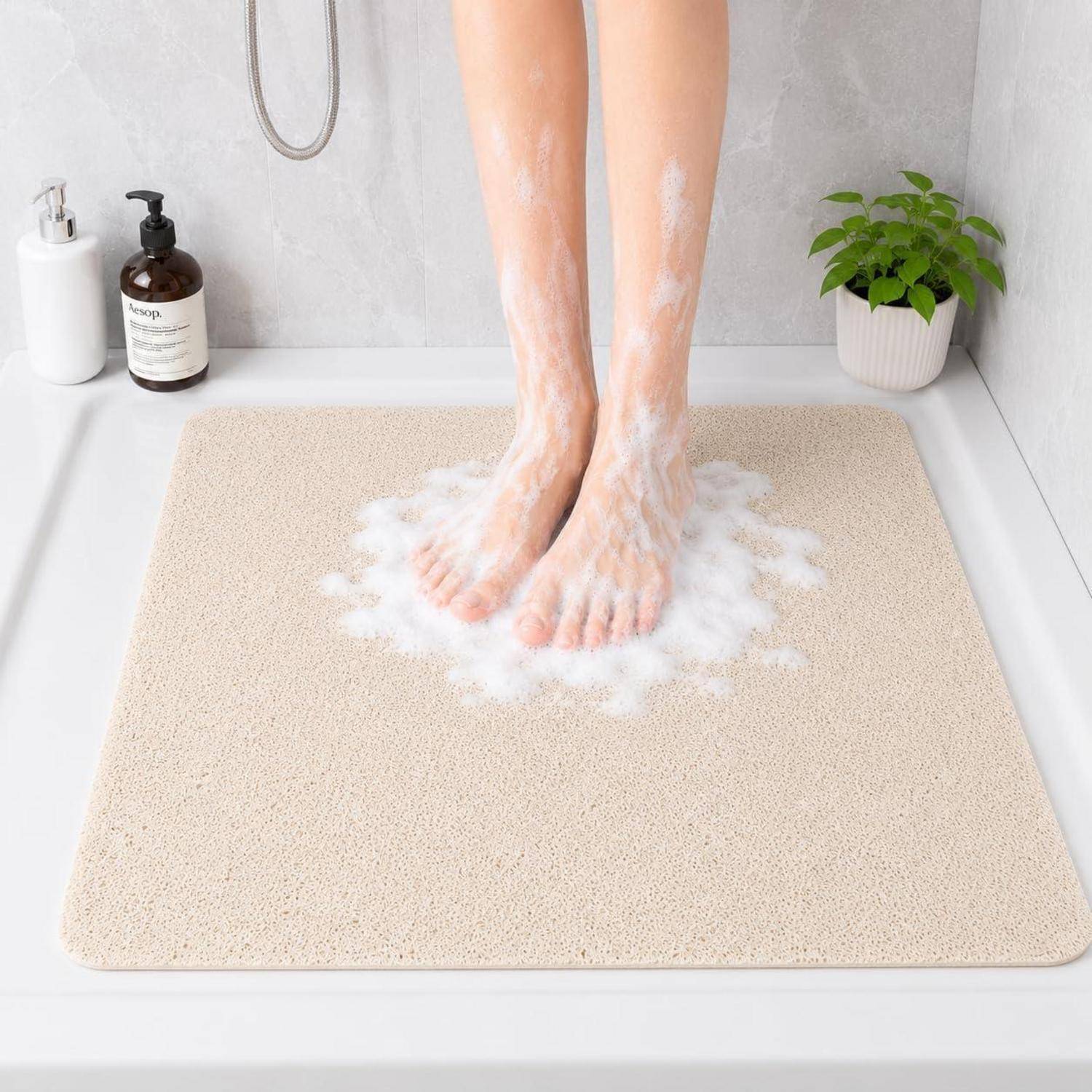 Eine Person steht auf einer beigen Badevorleger in einer Dusche und wäscht ihre Füße mit Seifenschaum; Toilettenartikel und eine Pflanze befinden sich in der Nähe.