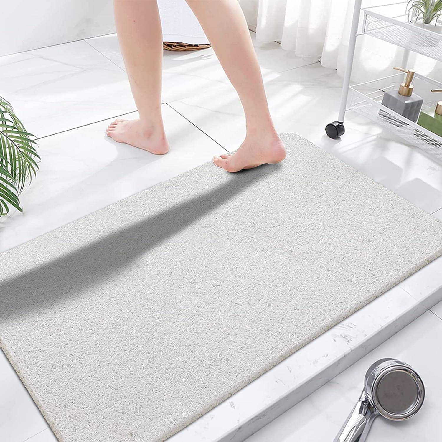 Die nackten Füße einer Person treten auf eine hellgraue Badematte in einem hellen Badezimmer mit weißen Fliesen und einem Duschkorb.
