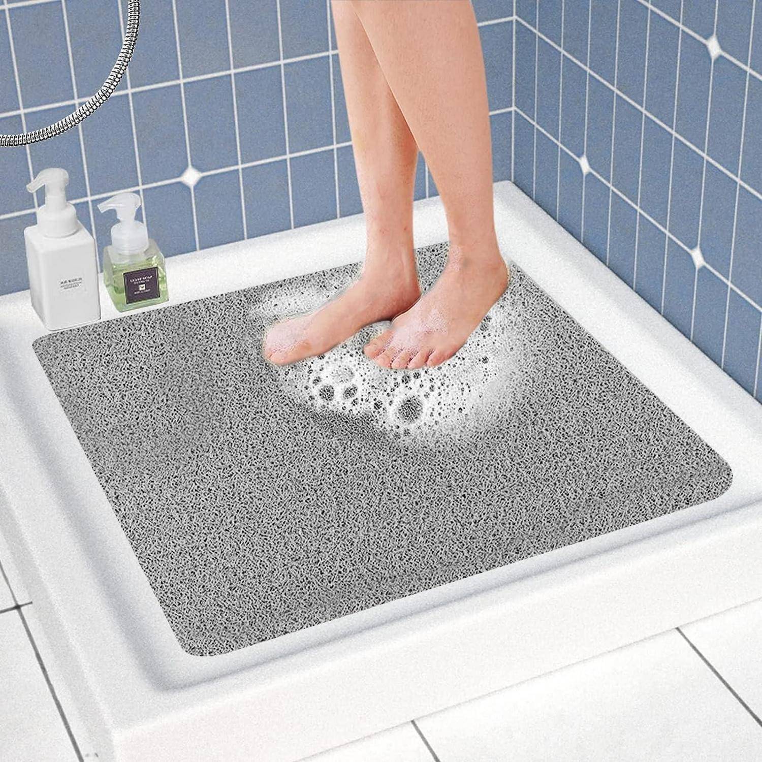 Füße stehen auf einer grauen rutschfesten Duschmatte in einer gefliesten Duschkabine mit Seife und Shampooflaschen in der Nähe.
