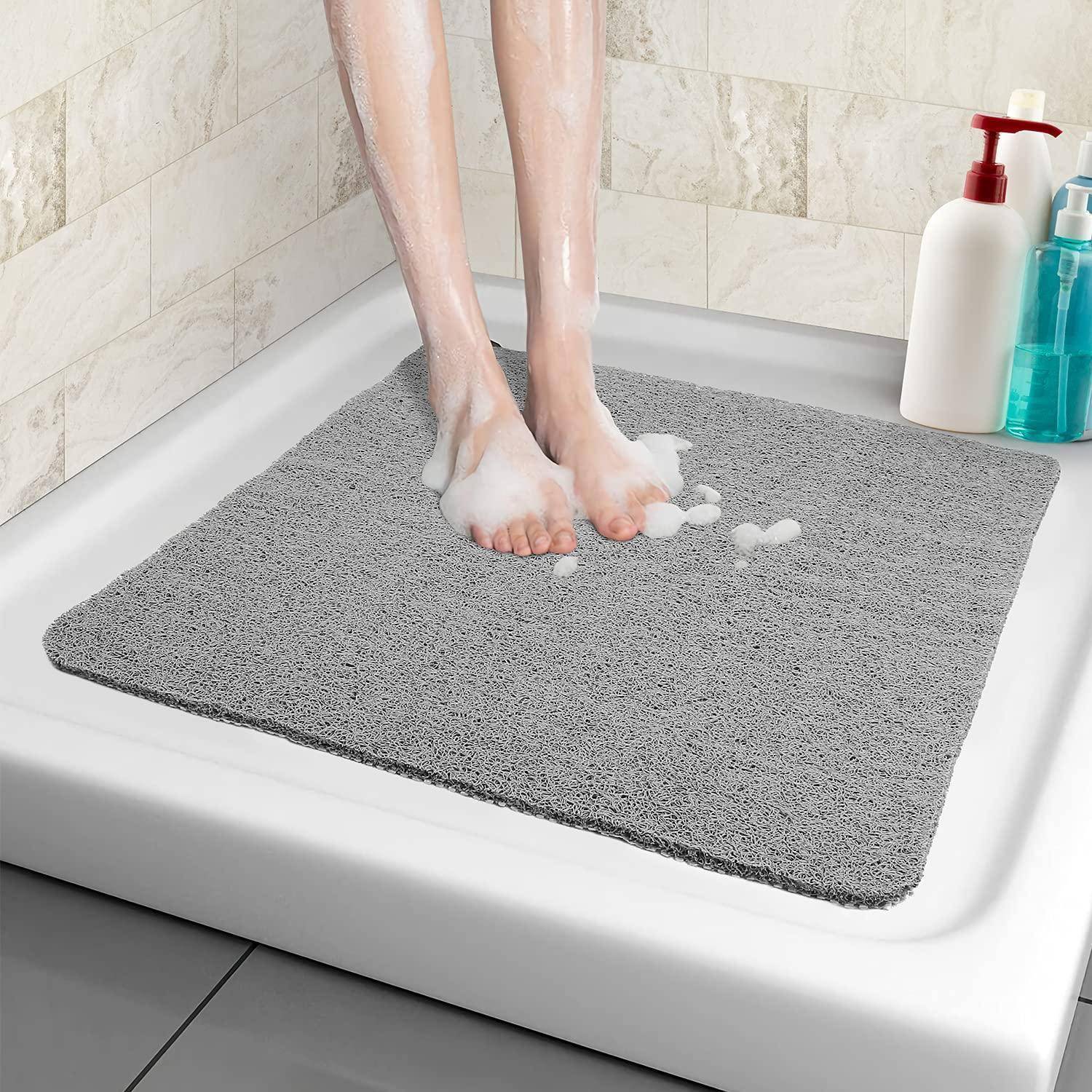Nackte Beine stehen auf einer grauen Badematte in einer Duschwanne, daneben stehen Seifen- und Shampooflaschen.