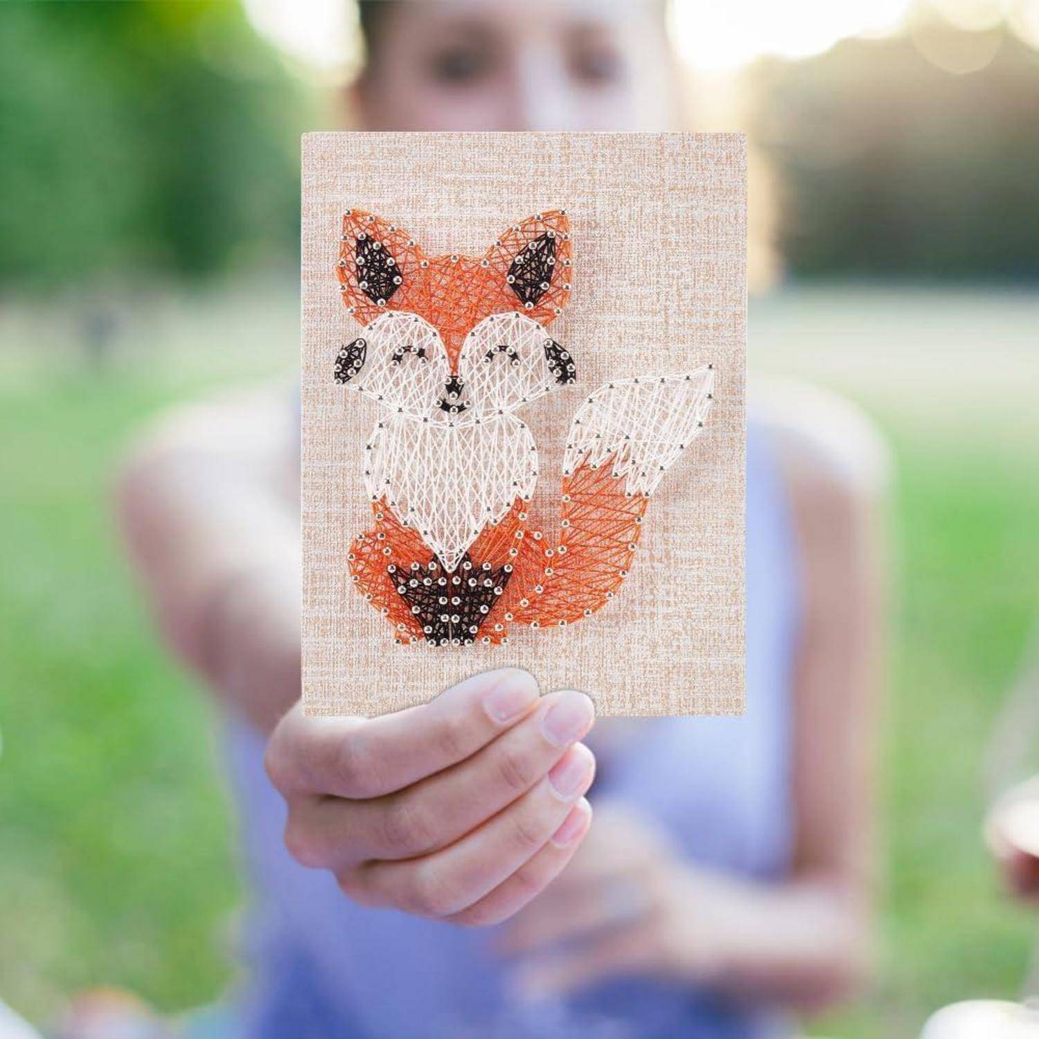 Eine Person hält eine handgefertigte Fuchsbastelkarte mit Fadenkunst und Perlen, die im Freien vor einem unscharfen grünen Hintergrund gezeigt wird.