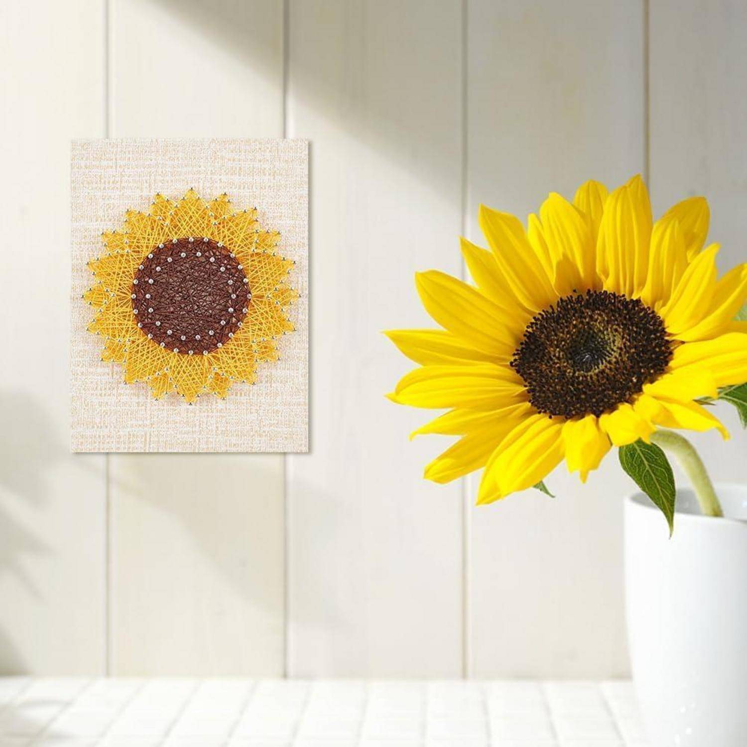 Leuchtend gelbe Sonnenblume in einer weißen Vase neben einem gerahmten Sonnenblumen-Kunstwerk an einer hellen Wand.