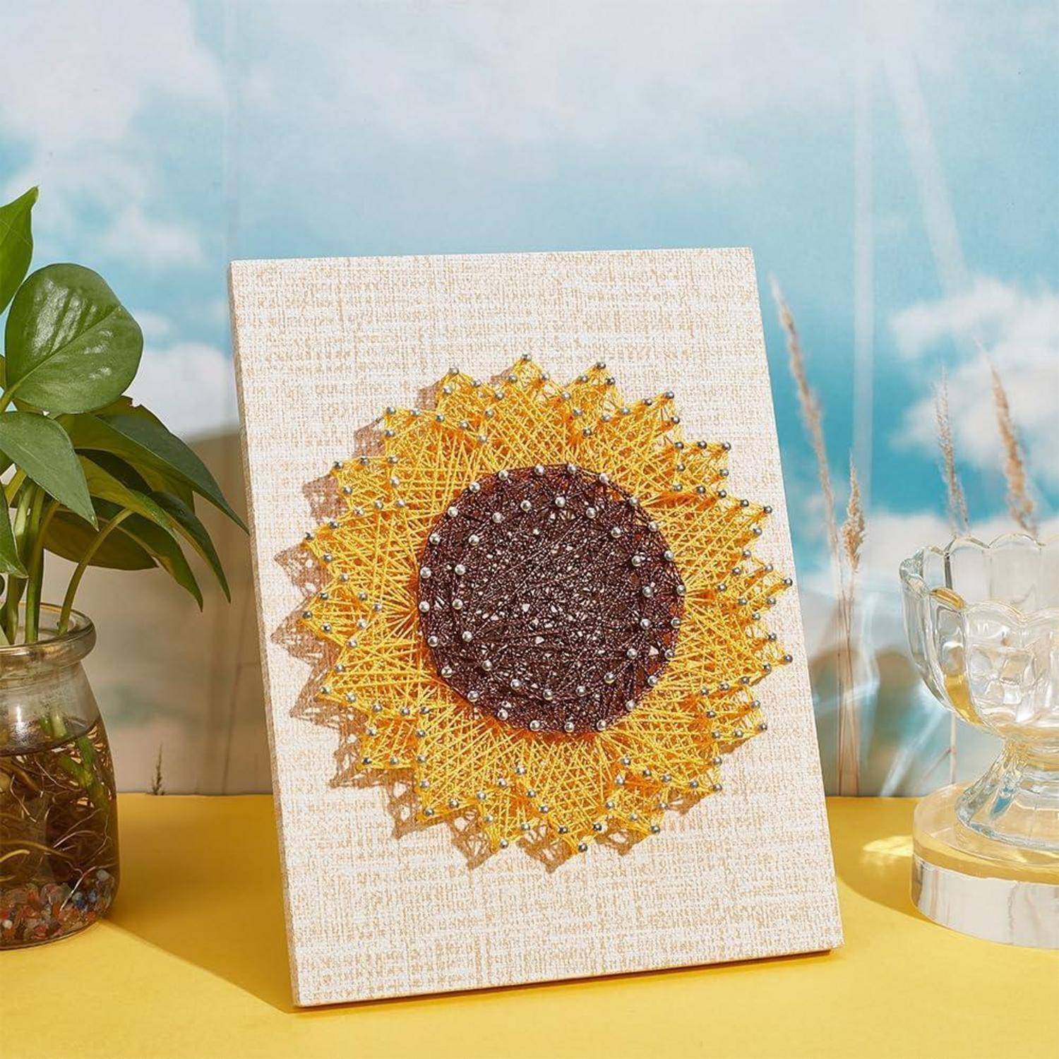 Dekorative Fadengrafik einer Sonnenblume auf einer Leinwand, mit gelben und braunen Fäden auf einer Tischplatte neben einer Pflanze und Glaselementen.