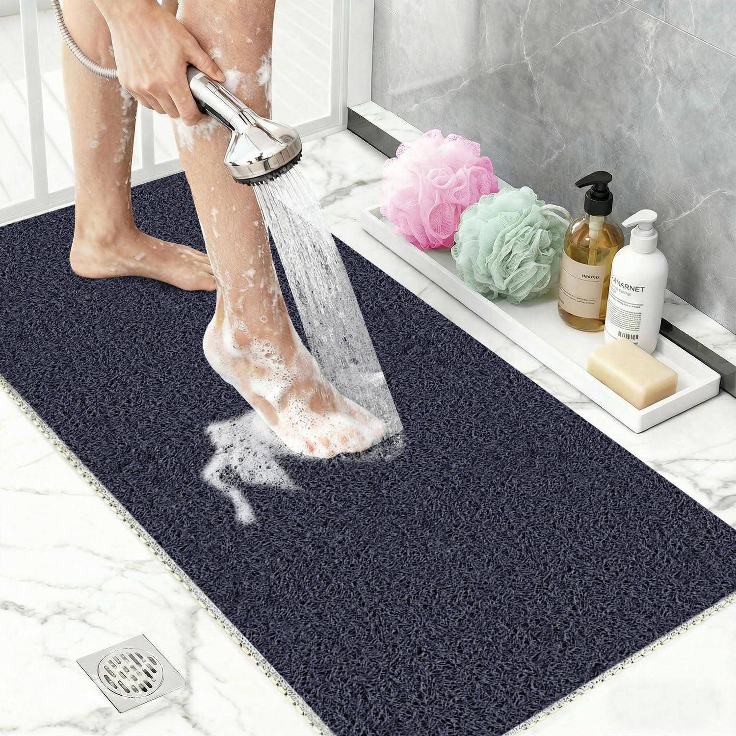 Eine Person spült eine dunkle strukturierte Badmatte unter der Dusche ab, während Badeschwämme und Toilettenartikel auf einem nahegelegenen Regal stehen.