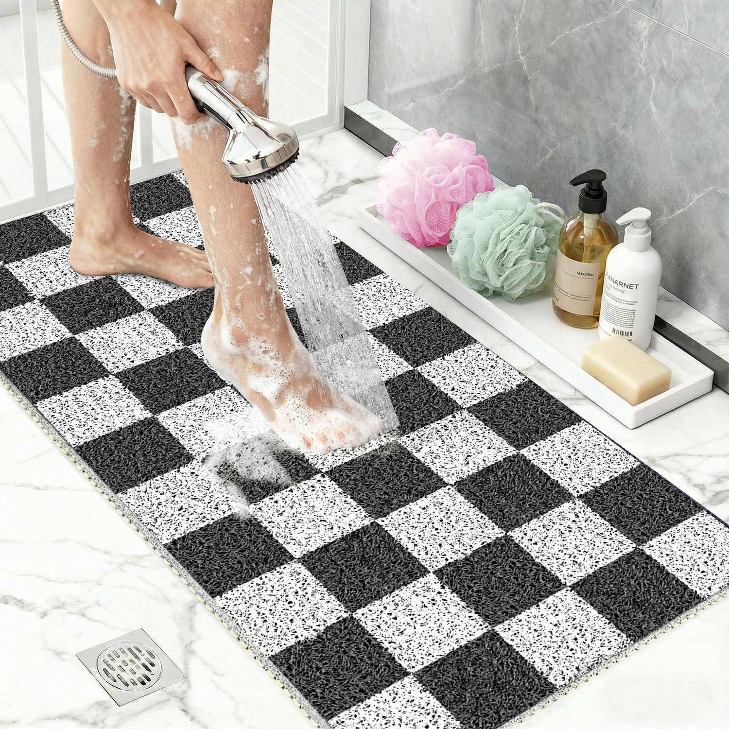 Person, die schaumige Füße in einer Dusche auf eine schwarz-weiße Badewannenmatte neben Badeprodukten und Luffas spült.
