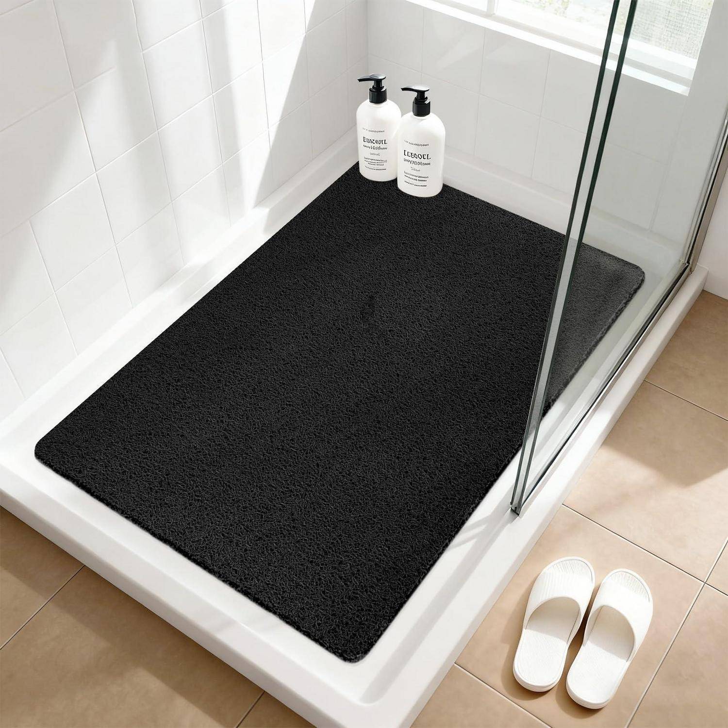 Schwarze Duschbadematte in einer ebenerdigen Dusche, mit zwei Flaschen auf der Ablage und weißen Hausschuhen draußen auf beigefarbener Fliese.