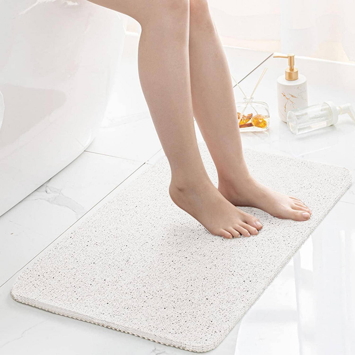 Barfüße, die auf einer weißen strukturierten Badematte in einem hellen Badezimmer stehen, mit einem Seifenspender und Handtüchern in der Nähe.