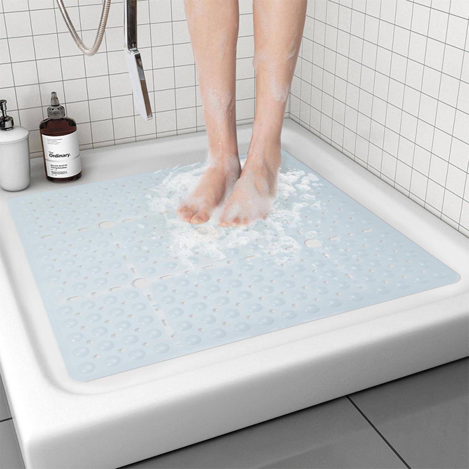 Person steht auf einer hellblauen rutschfesten Badewannenmatte unter der Dusche, mit Wasser und Seifenschaum auf der Matte neben einer gefliesten Wand und Badeprodukten.