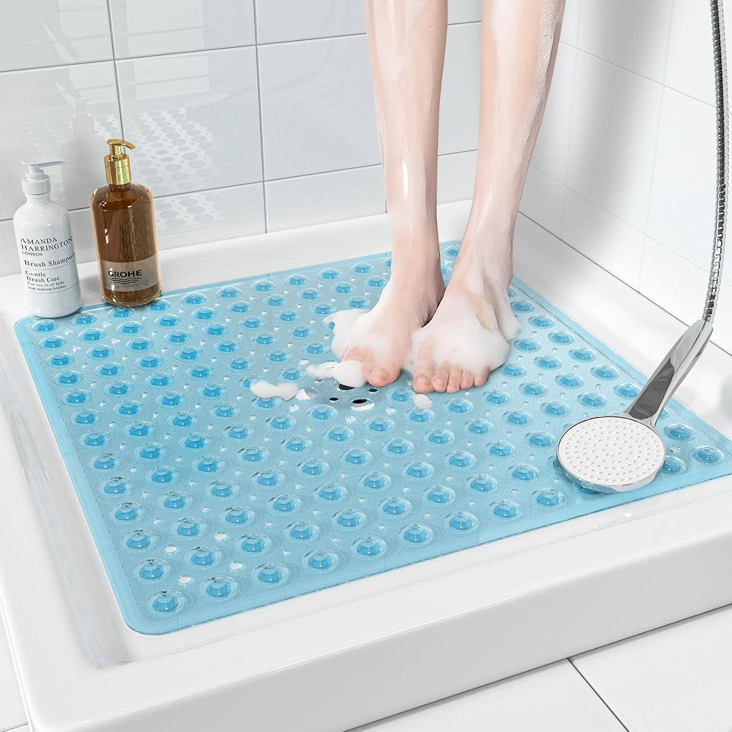 Füße stehen auf einer blauen rutschfesten Badematte in einer mit weißen Kacheln gefliesten Dusche, mit Seifenflaschen und einem Duschkopf in der Nähe.