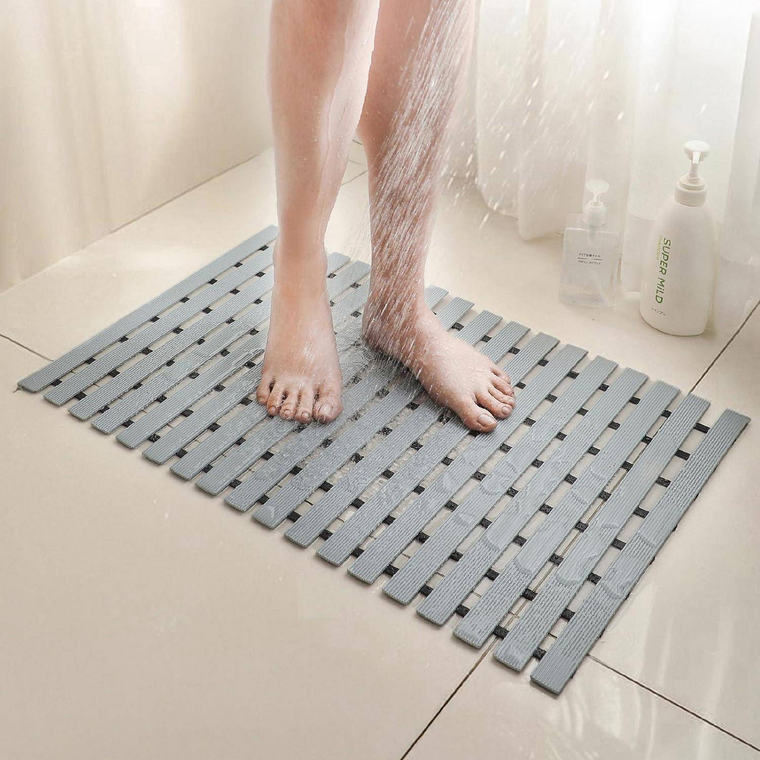 Eine Person duscht auf einer grauen Duschmatte mit Lamellen in einem gefliesten Badezimmer, wobei Wasser über ihre Füße spritzt und Shampooflaschen in der Nähe stehen.
