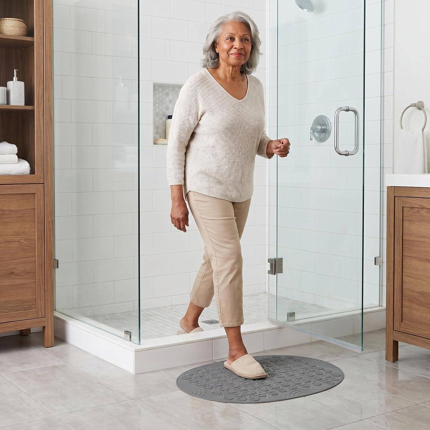 Eine ältere Frau, die aus einer gläsernen Dusche in einem hellen Badezimmer heraustritt, hält die Türklinke fest und trägt Pantoffeln auf einer Badematte.