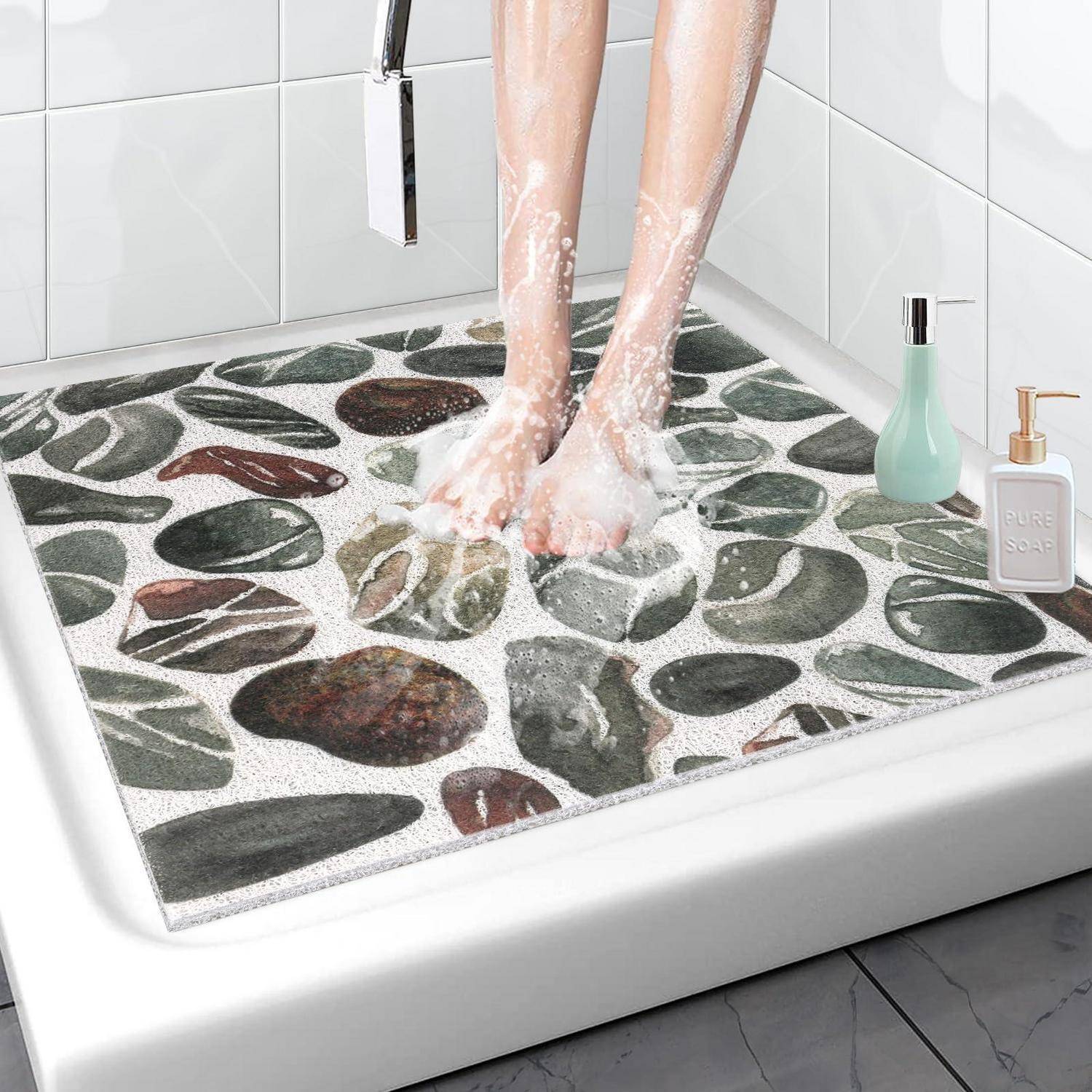 Eine Person steht auf einer Badematte mit Kieselsteinmuster in einer Dusche, mit Seifenschaum an den Füßen und Toilettenartikelfläschchen in der Nähe.