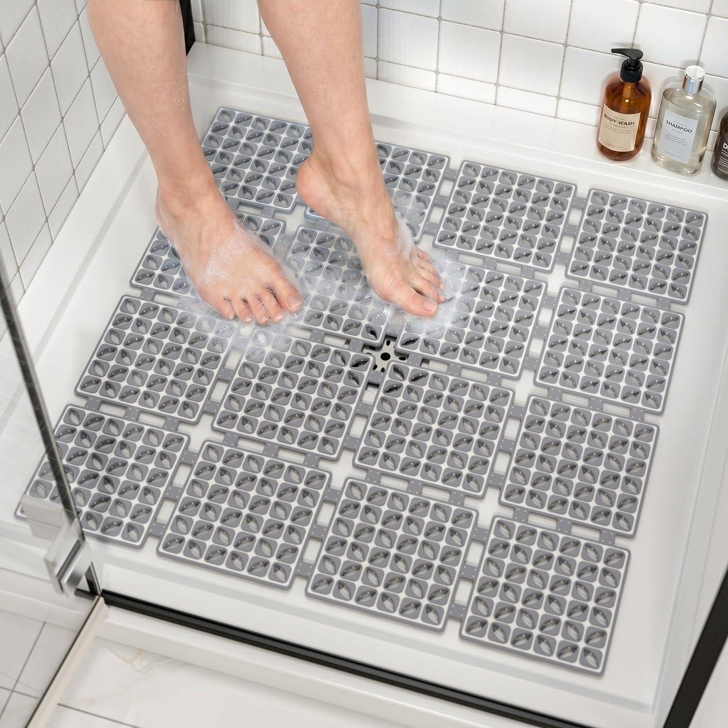 Nackte Füße, die auf einem Duschboden mit vielen grauen Antirutsch-Saugmatten stehen, mit Seifenflaschen auf dem Regal.