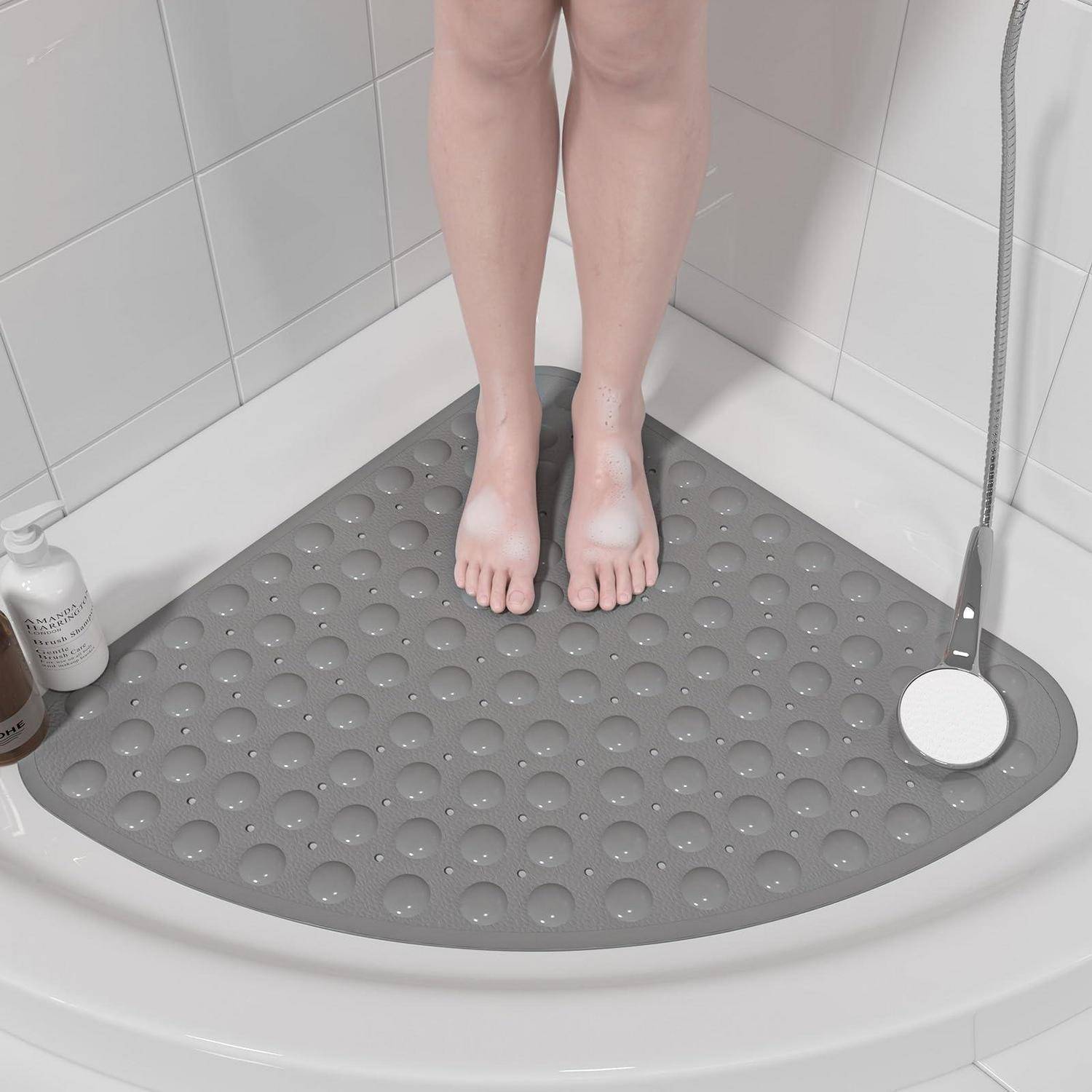 Person stands barefoot on a gray non-slip shower mat in a tiled shower stall, with a handheld shower and bath products nearby.