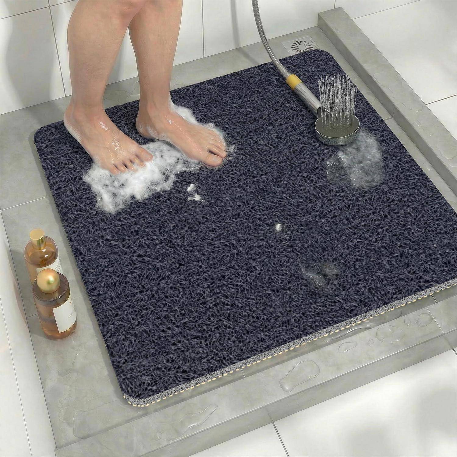 Person duscht, während sie auf einer dunklen Badematte im Badezimmer steht, mit Wasser, das aus einer Handbrause spritzt, und Seifenflaschen in der Nähe.