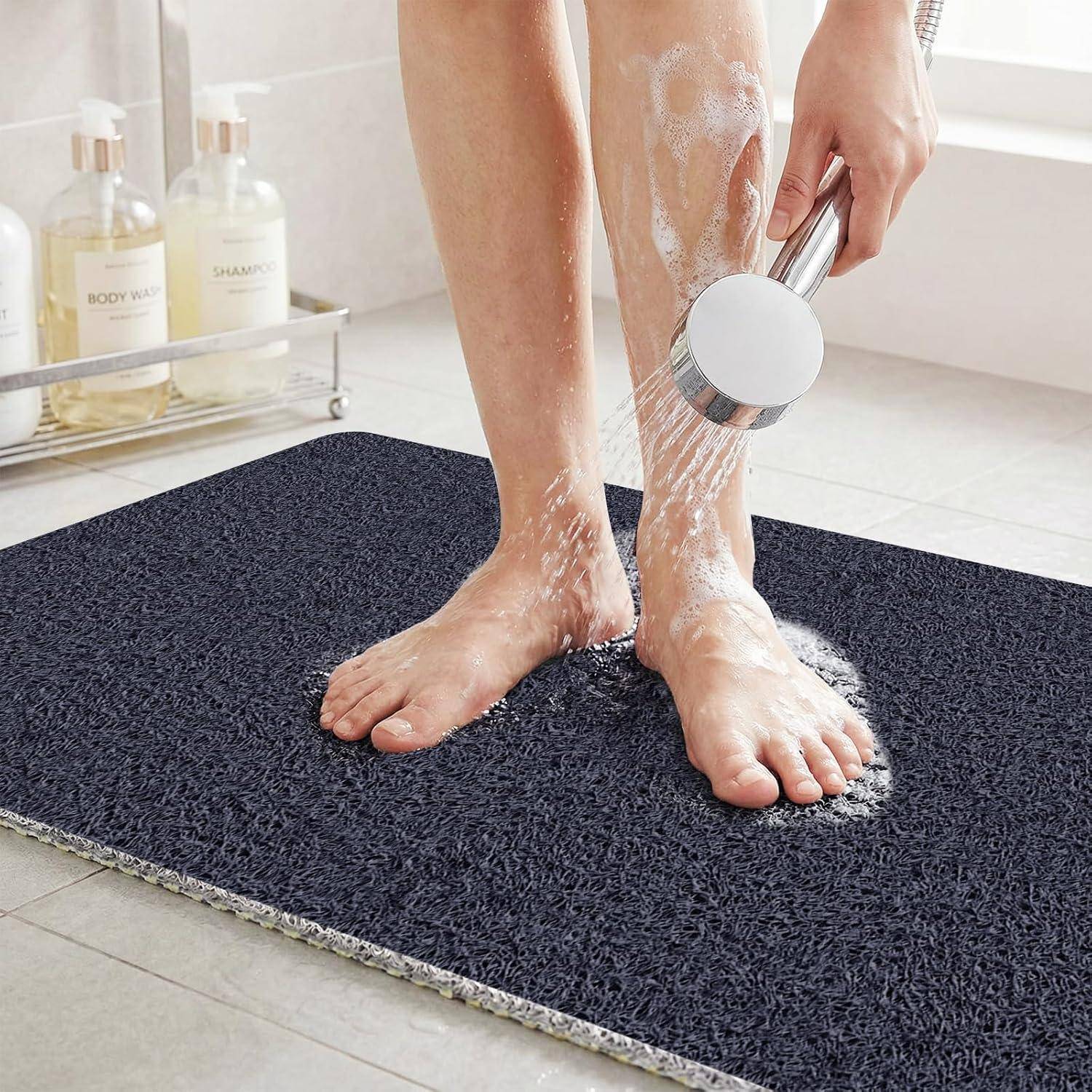 Person spült Seife von den Füßen mit einer Handbrause über einer dunklen Badezimmermatte in einem Duschbereich ab.