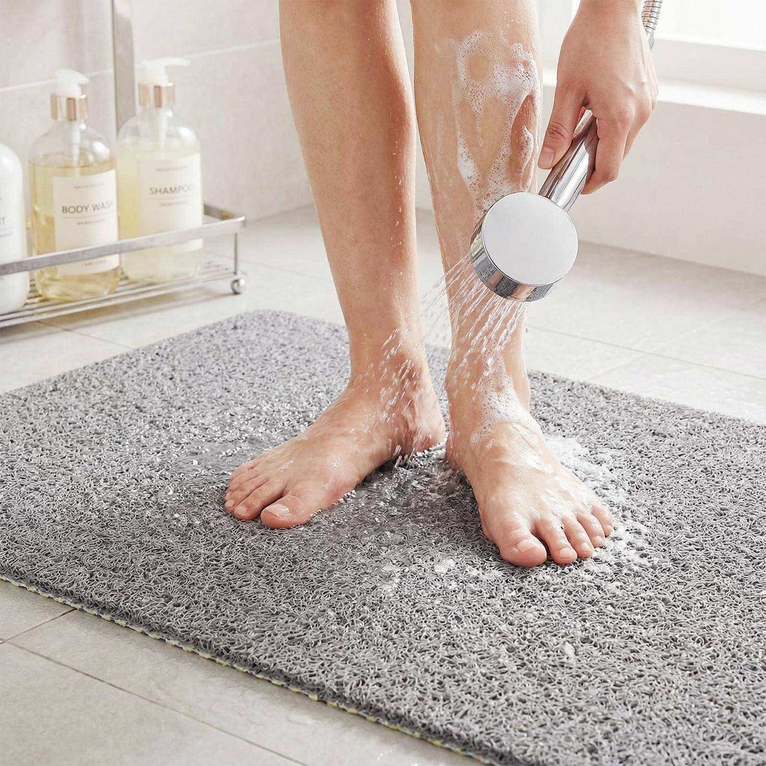Eine Person spült ihre Beine und Füße mit einer Handbrause über einer grauen Badmatte im Badezimmer.