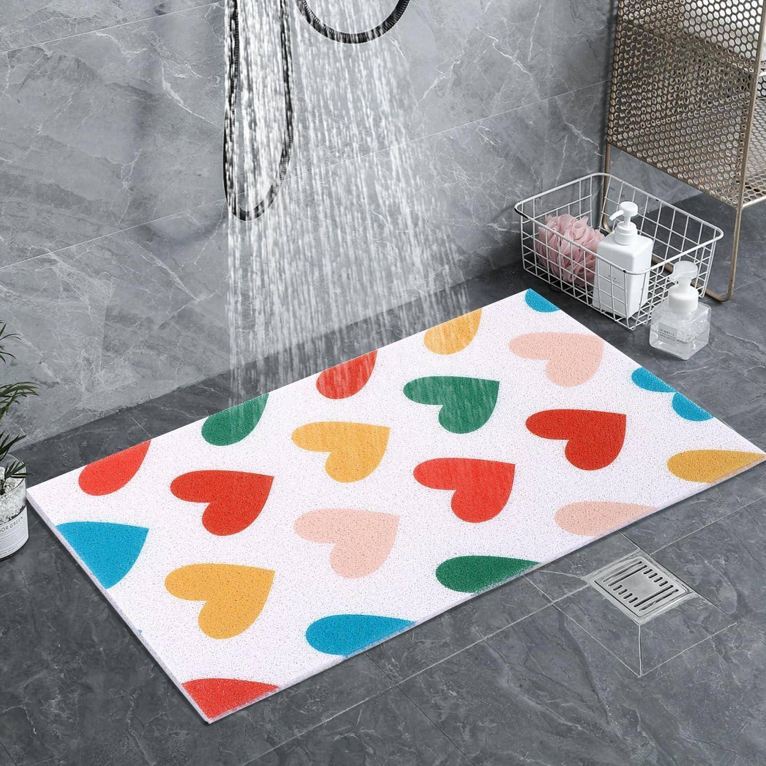 Bunte herzförmige Badematte auf einem grauen Duschtablett unter fließendem Wasser, mit Toilettenartikeln in einem Drahtkorb in der Nähe.