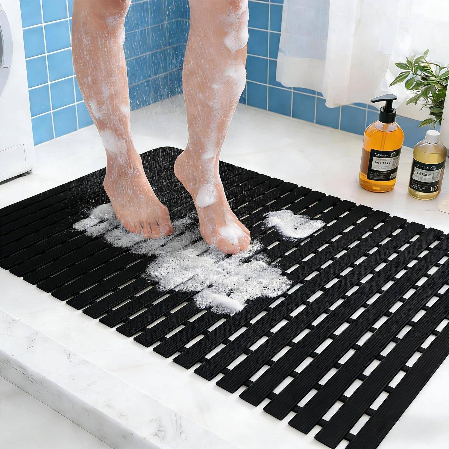 Person spült sich seifige Füße auf einer schwarzen Badematte mit Lamellen in einer gekachelten Dusche ab, in der Nähe von Shampooflaschen.