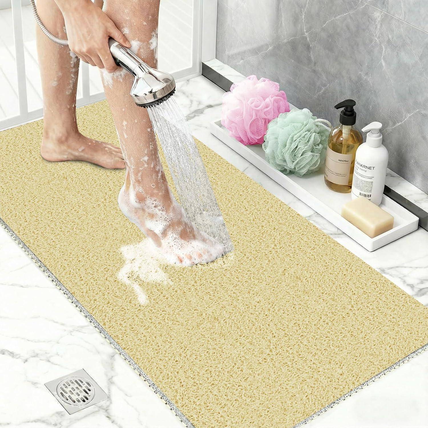 Person, die schaumige Füße auf einer strukturierten gelben Badematte in einer Dusche abspült, mit Badeschwämmen und Flaschen auf einem Regal in der Nähe.