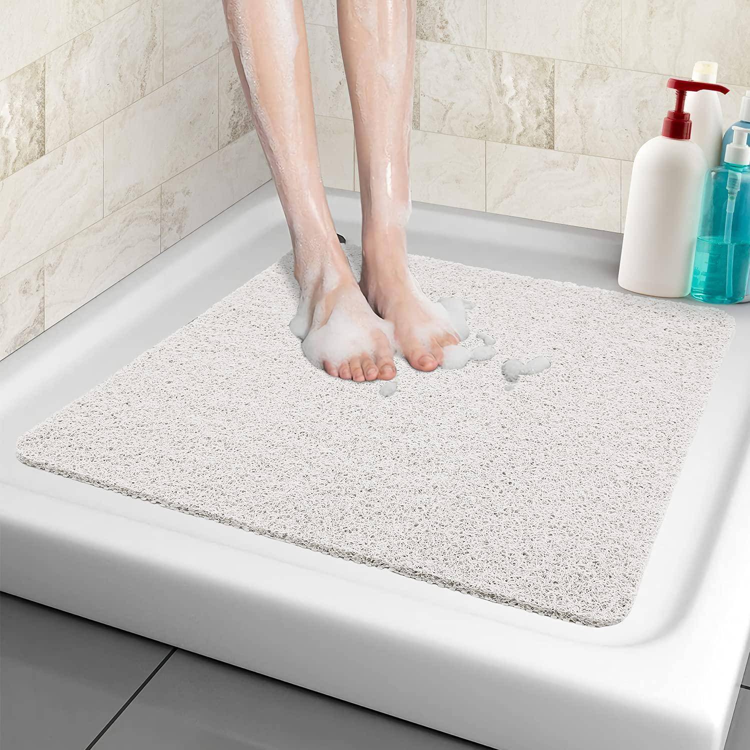 Person, die sich auf einer weißen strukturierten Badematte in einer Duschkabine die Füße wäscht, mit Seifenschaum und Shampooflaschen in der Nähe.