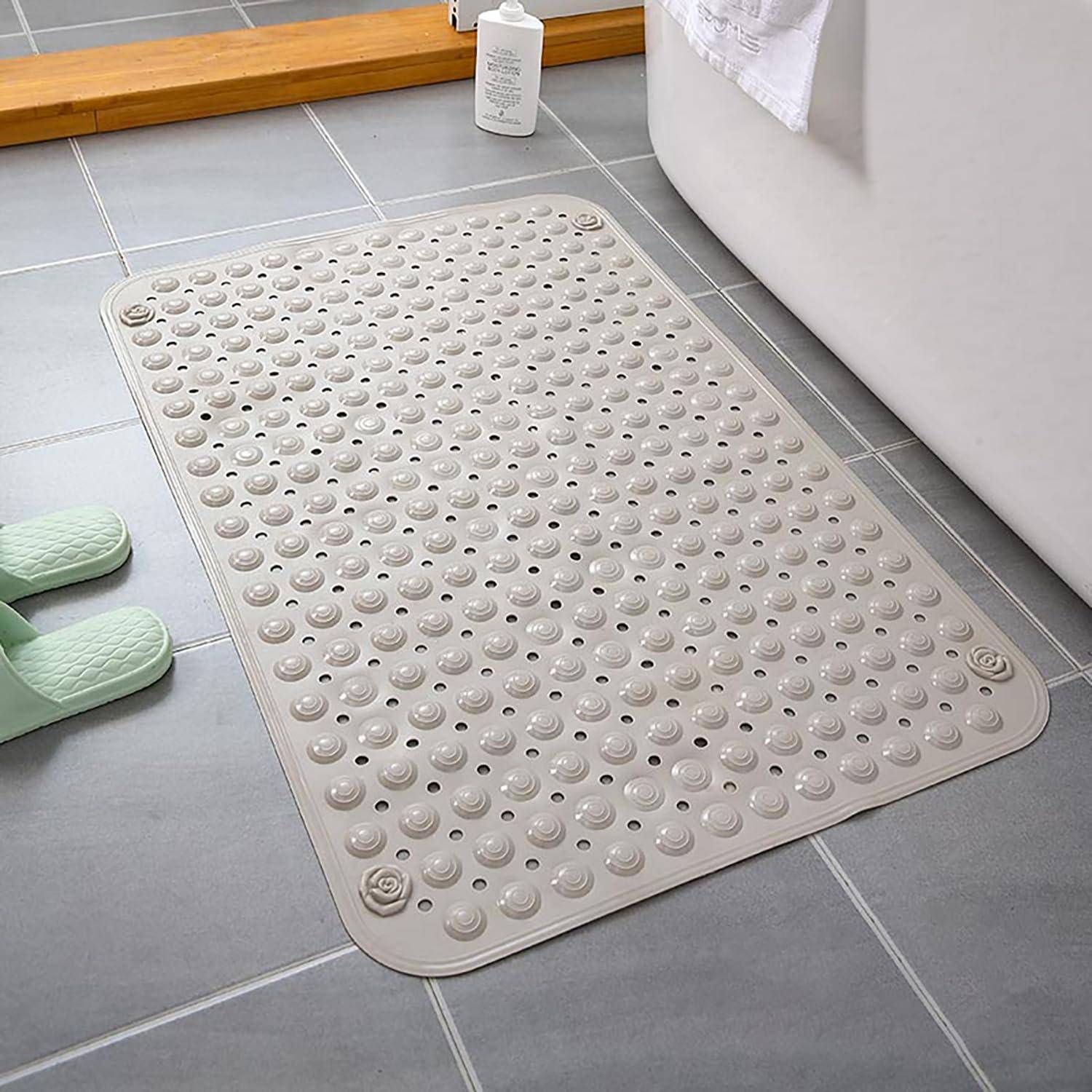 Beige strukturierte Badmatte mit saugnapfähnlichen Noppen auf einem grauen Fliesenbadezimmerboden neben einer Badewanne und Hausschuhen.