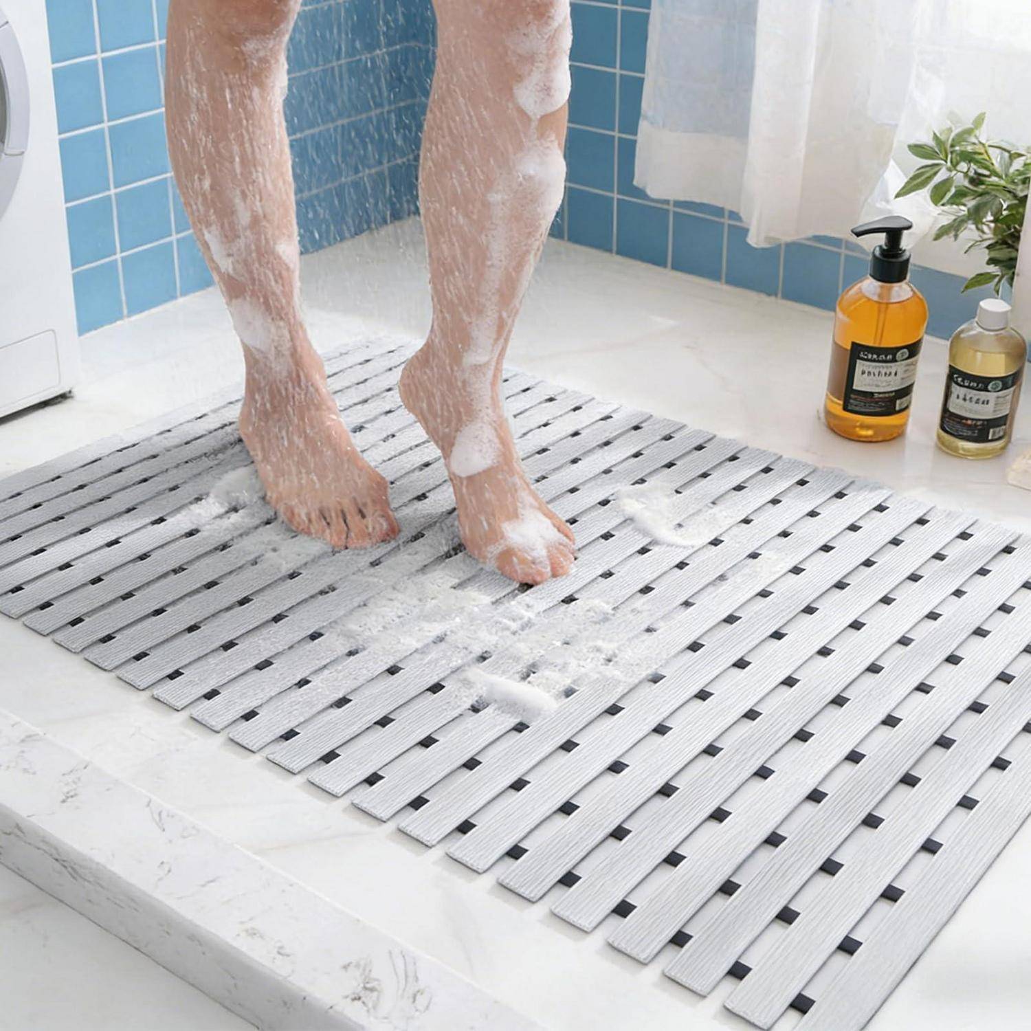 Eine Person, die barfuß auf einer Lamellenbadematte in einer Dusche steht, mit Seifenschaum an ihren Beinen und dem Boden, neben Shampooflaschen.