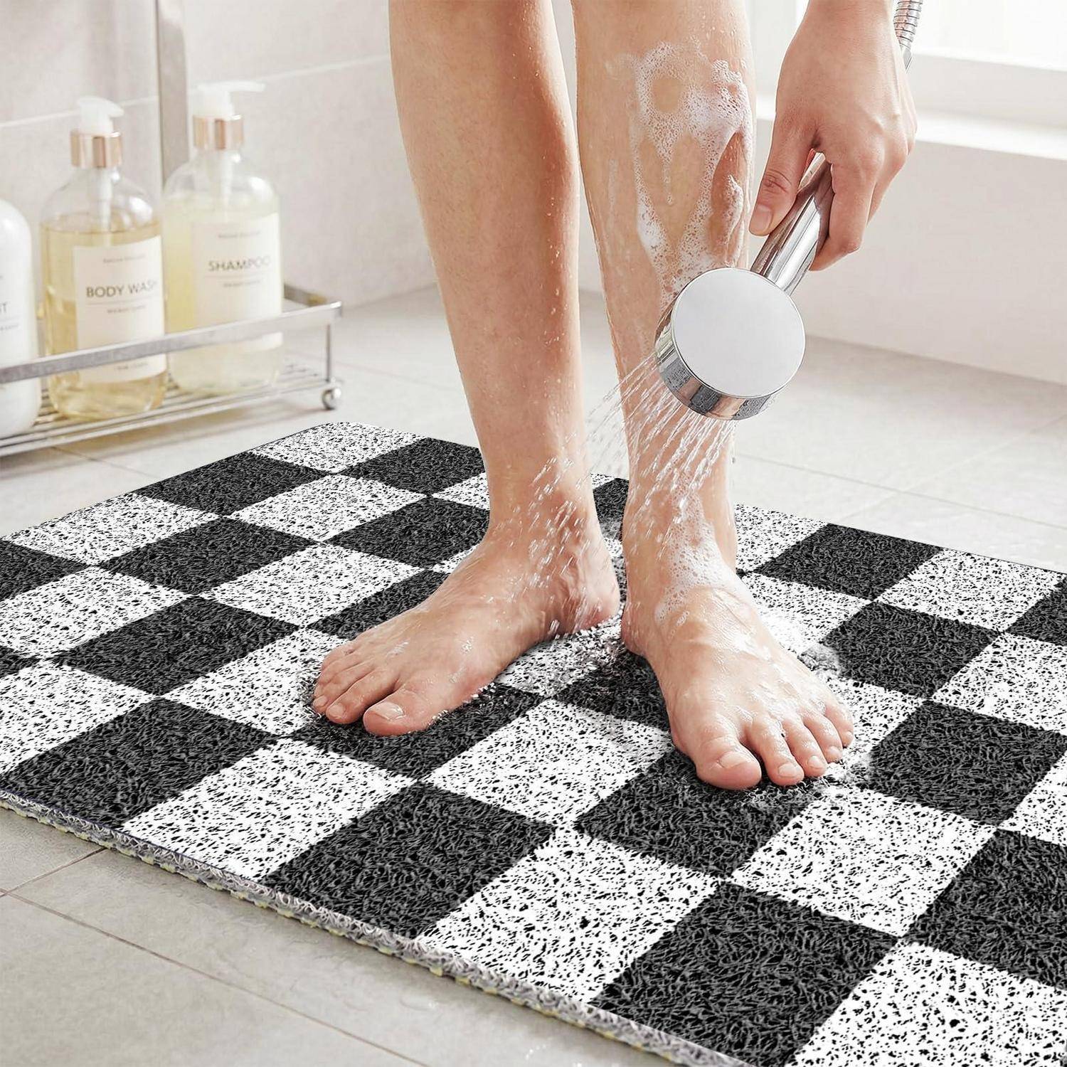 Eine Person spült ihre Beine und Füße mit einer Handbrause über einer schwarz-weißen karierten Badezimmermatte im Badezimmer ab.