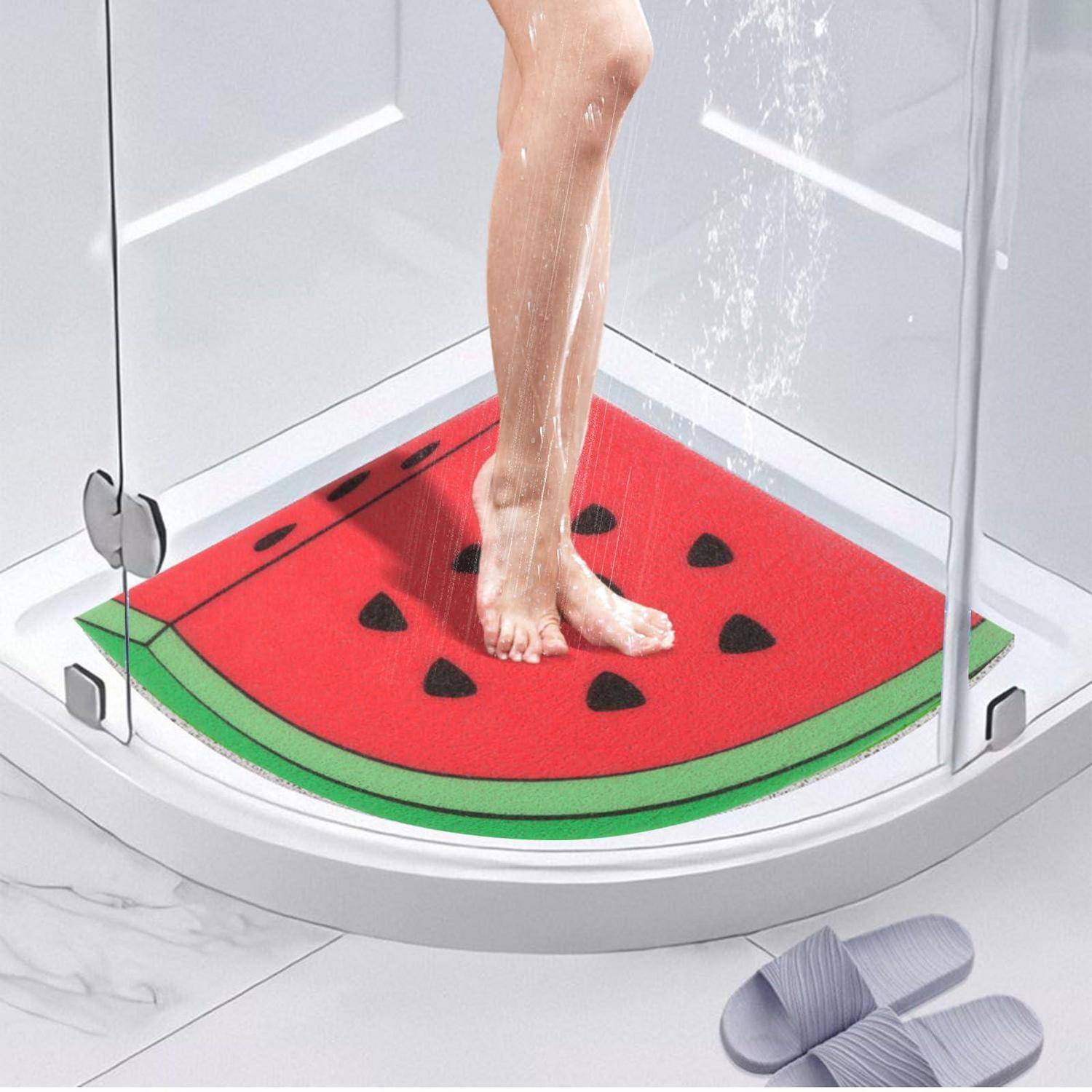Person, der/die unter der Dusche in einer Glaskabine steht, auf einer Badmatte mit Wassermelonen-Motiv stehend, mit Sandalen in der Nähe.
