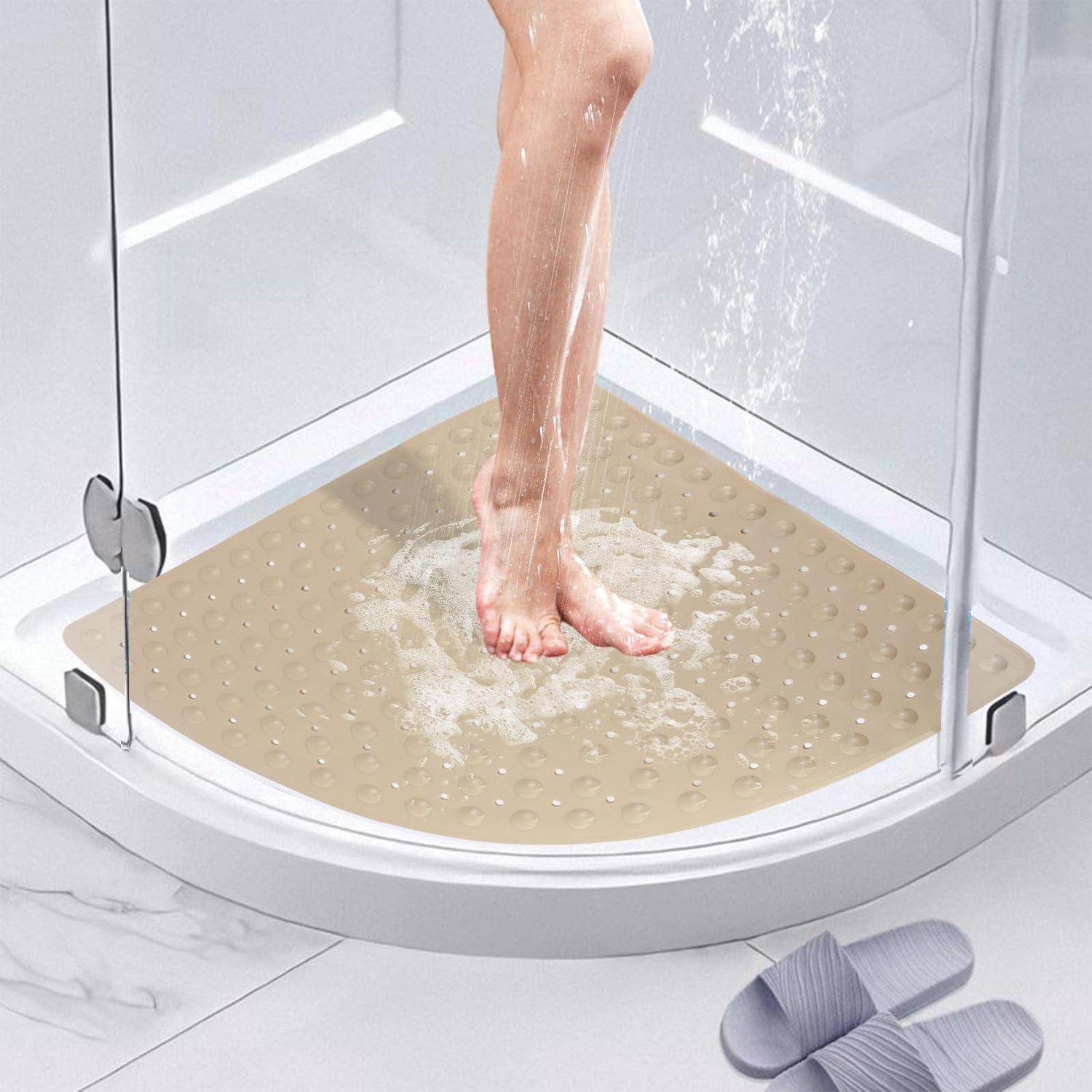 Nackte Beine stehen unter einer laufenden Dusche auf einer beigen rutschfesten Matte in einer Glasumrandung; Pantoffeln ruhen außerhalb der Dusche.
