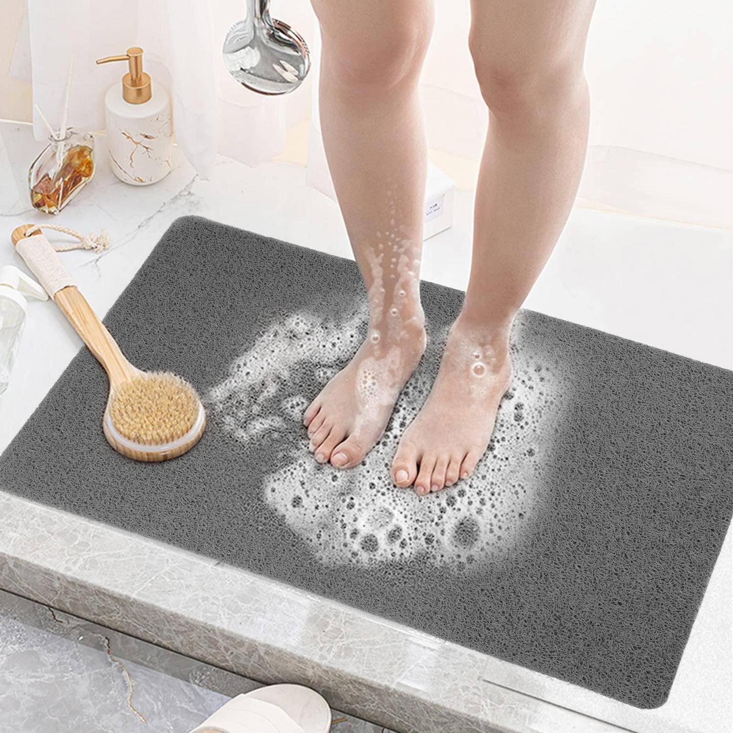Person, die seifige Füße auf einer grauen Badmatte neben einer Badewanne ausspült, mit einem Duschkopf und einer Badebürste in der Nähe.