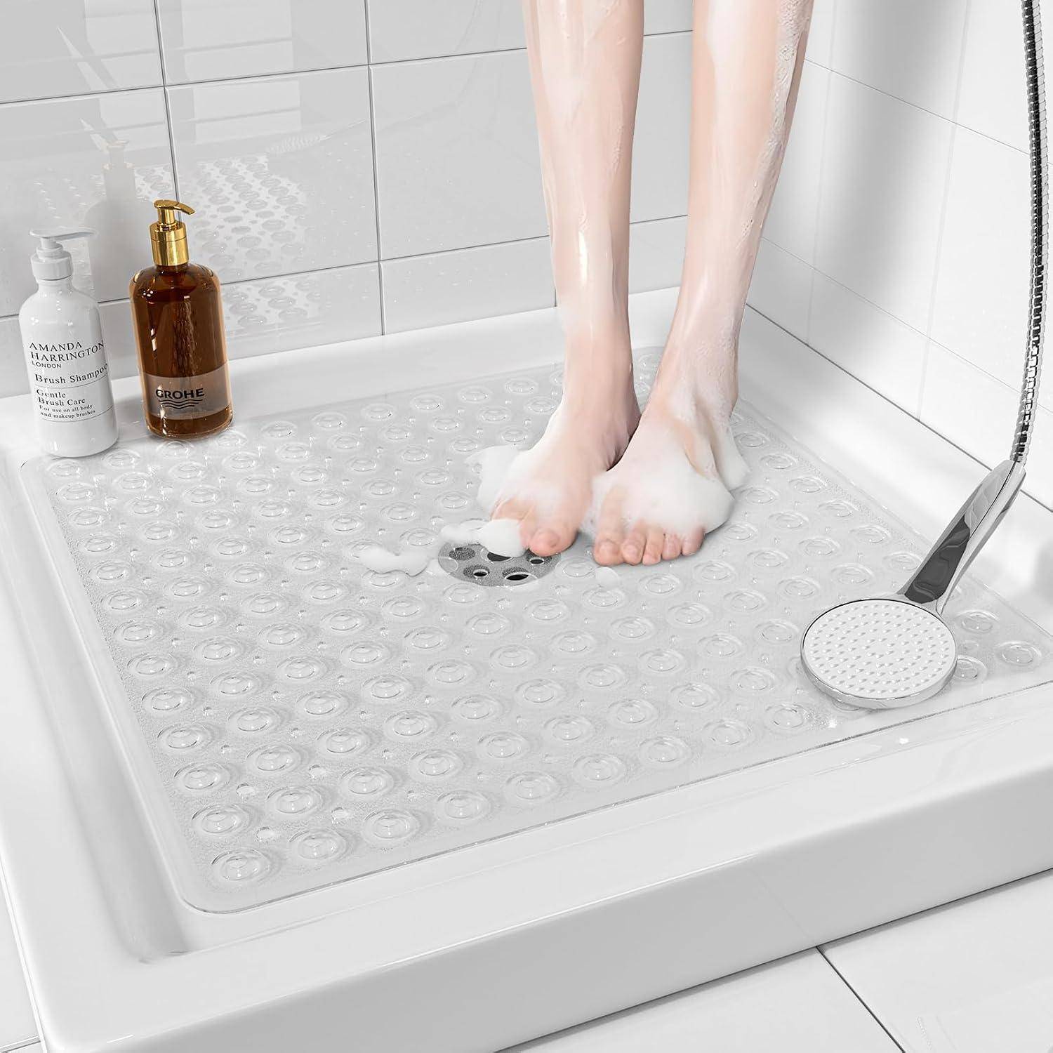 Füße stehen auf einer durchsichtigen Duscheinlage in einer weiß gefliesten Dusche, mit Shampooflaschen und einem Handbrausekopf in der Nähe.