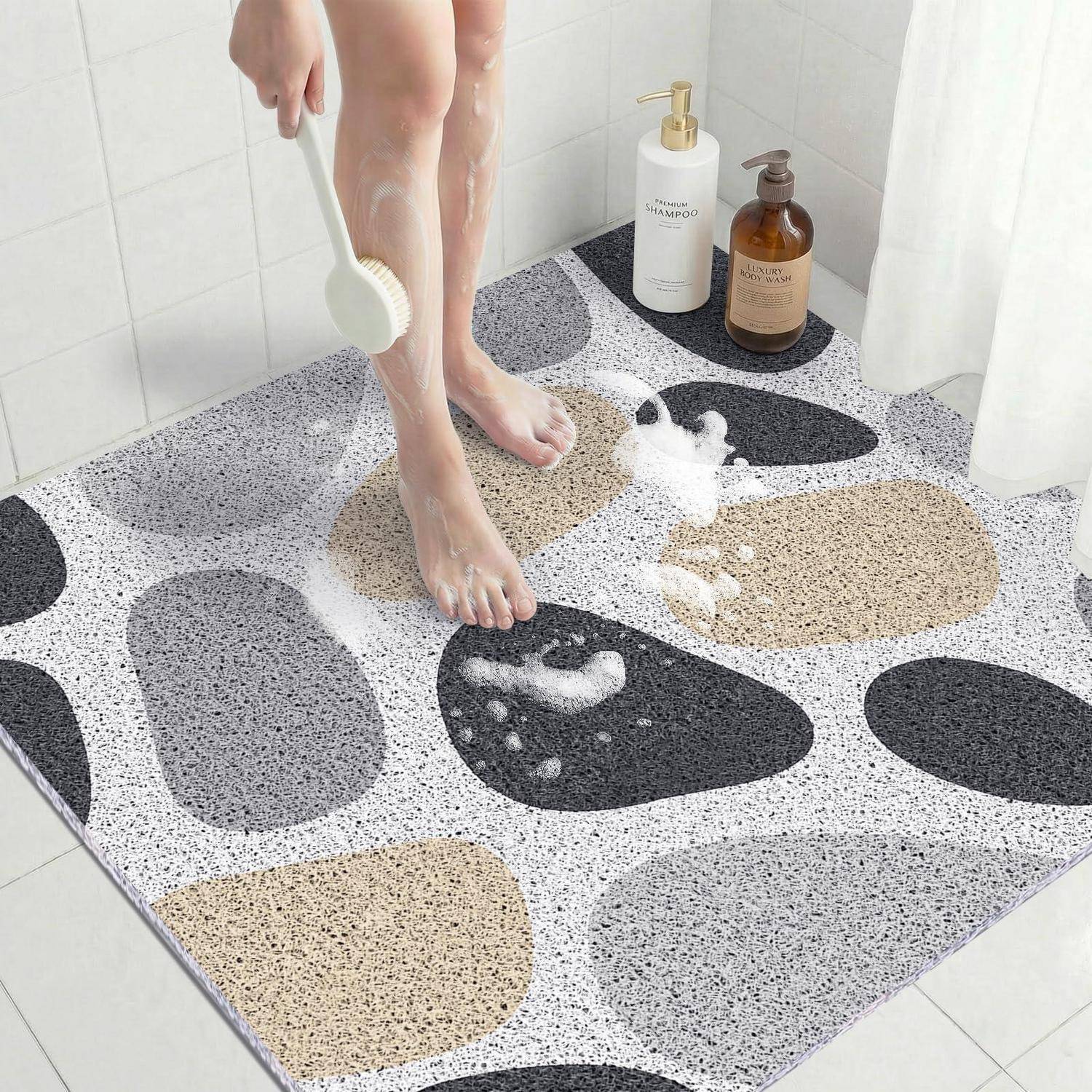 Eine Person spült ihre seifigen Füße in einer Dusche auf einer Badematte mit Kieselmuster ab, mit Shampooflaschen neben dem Duschvorhang.