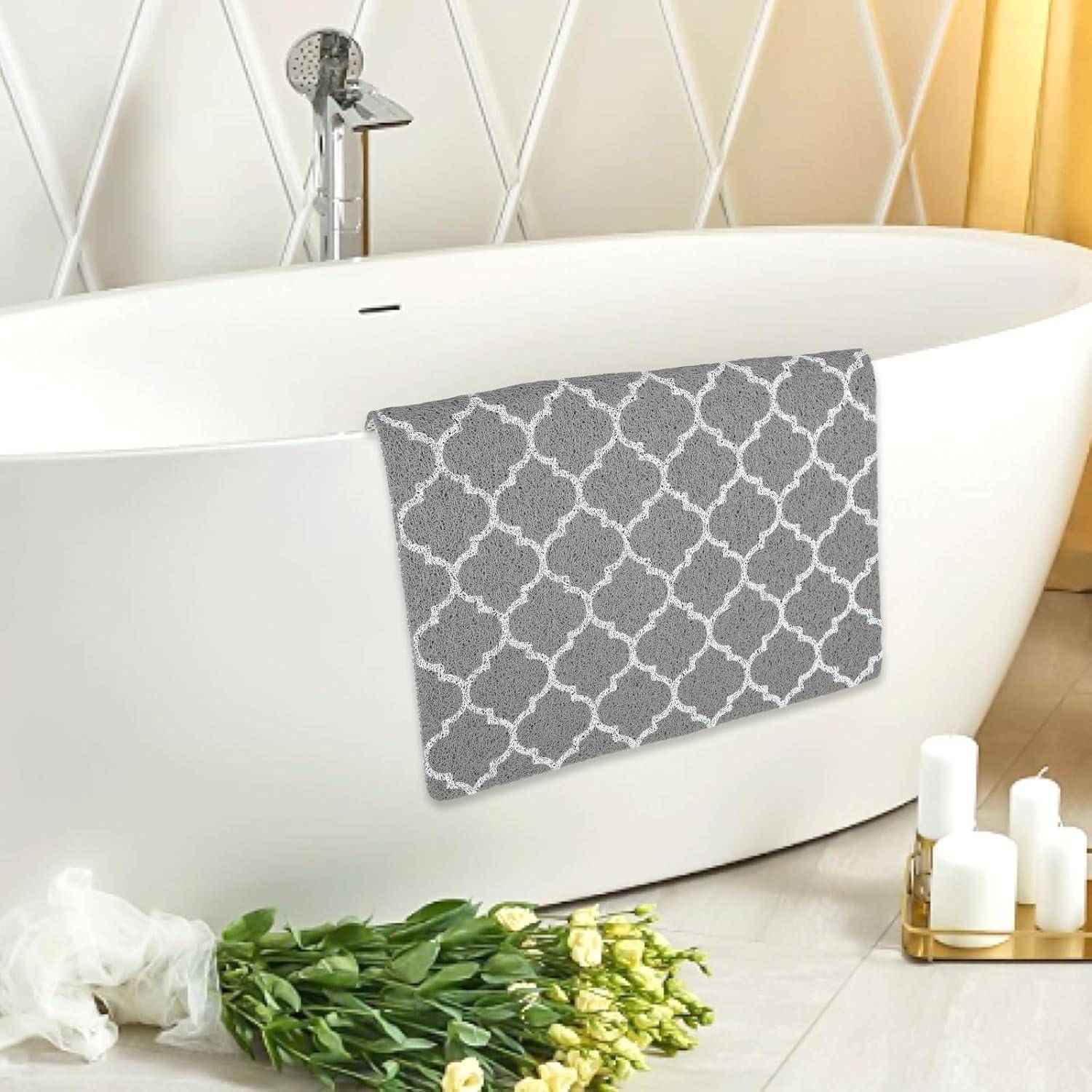 Freistehende weiße Badewanne mit grauem gemustertem Badteppich, der über die Seite drapiert ist, frische Blumen und Kerzen daneben in einem hellen Badezimmer.