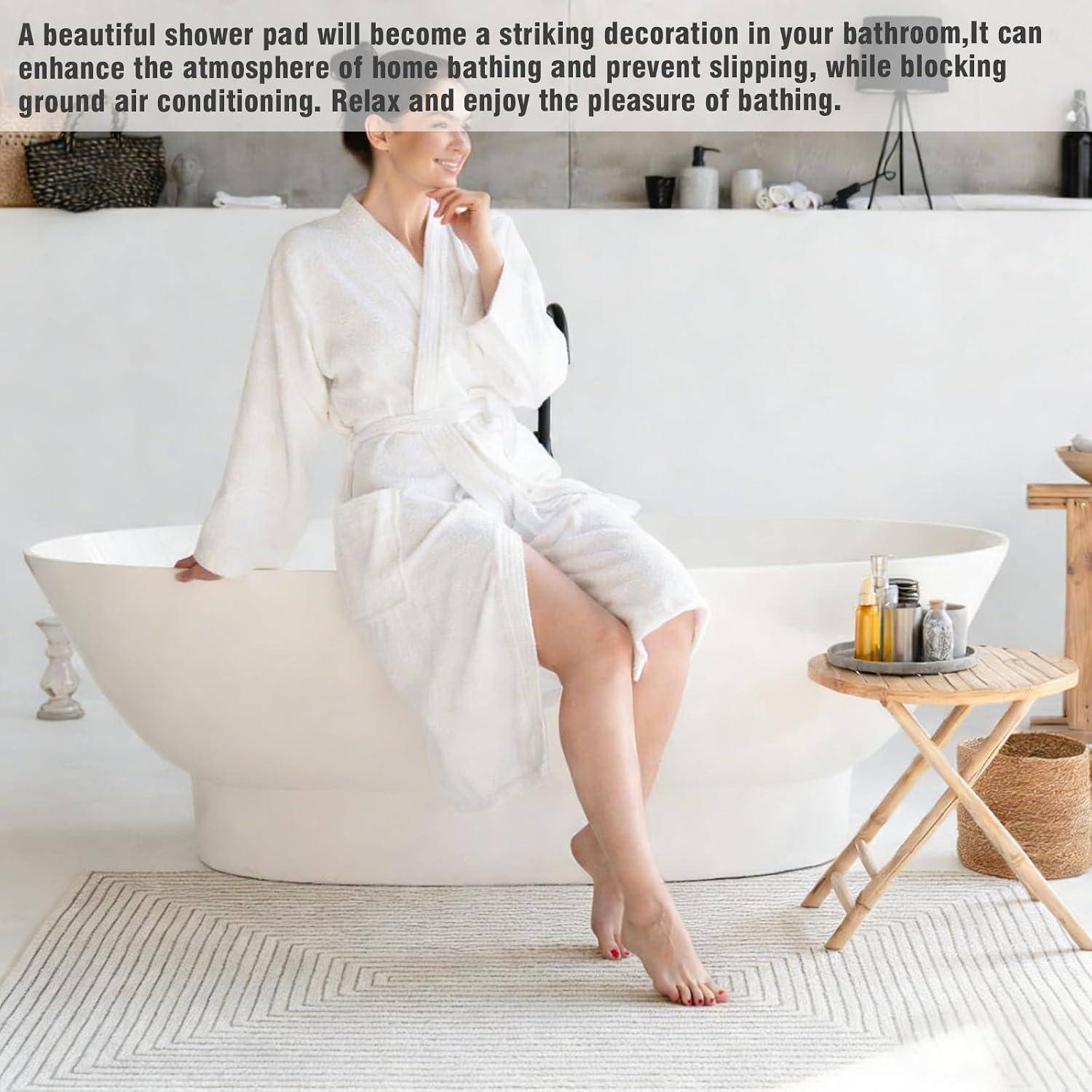 Eine Frau in einem weißen Bademantel sitzt auf einer freistehenden Badewanne in einem hellen Badezimmer, mit einer Badematte im Vordergrund und Badaccessoires in der Nähe.