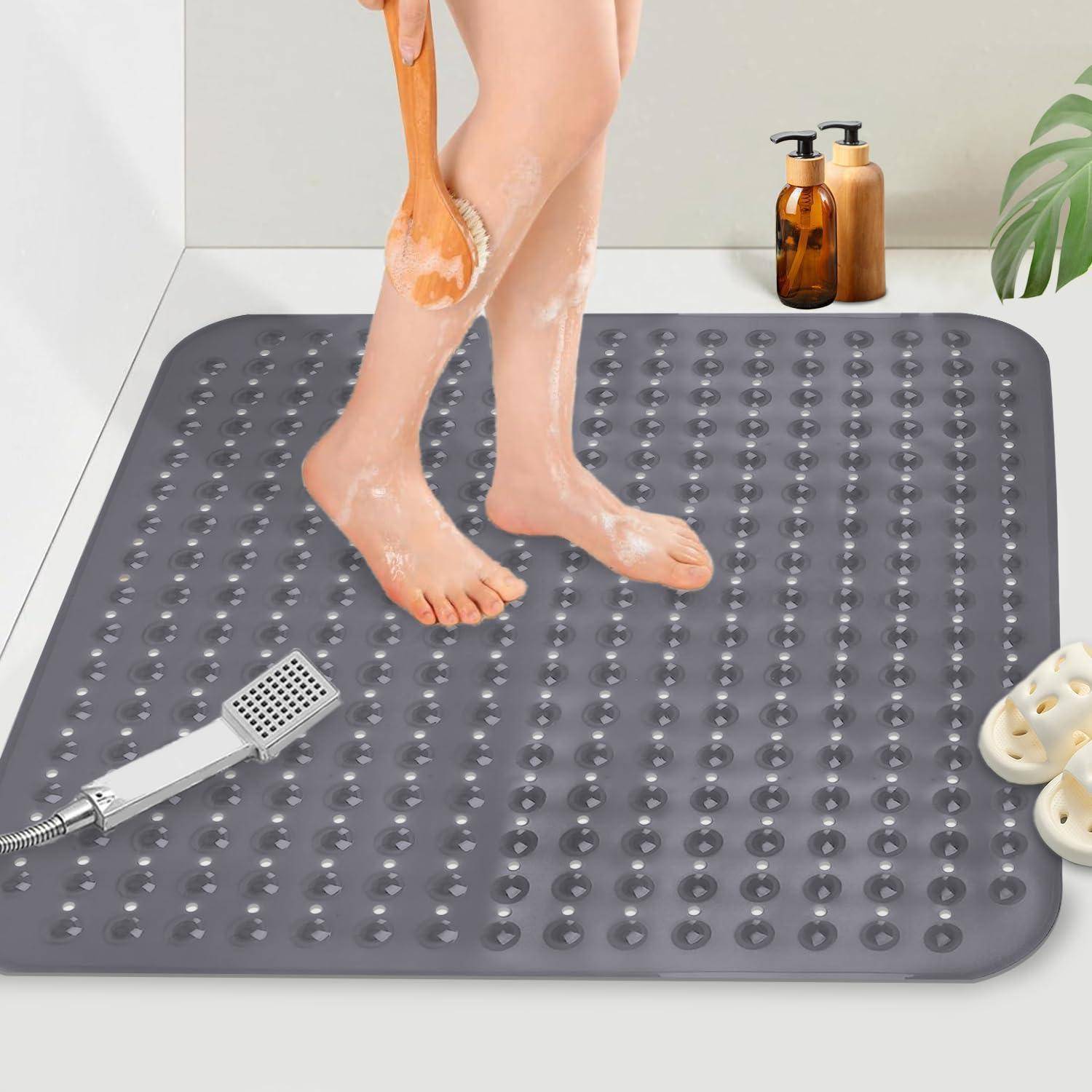 Person, die auf einer grauen rutschfesten Badematte duscht, während sie ihr Bein mit einer Bürste schrubbt; Handbrause und Toilettenartikel befinden sich in der Nähe.